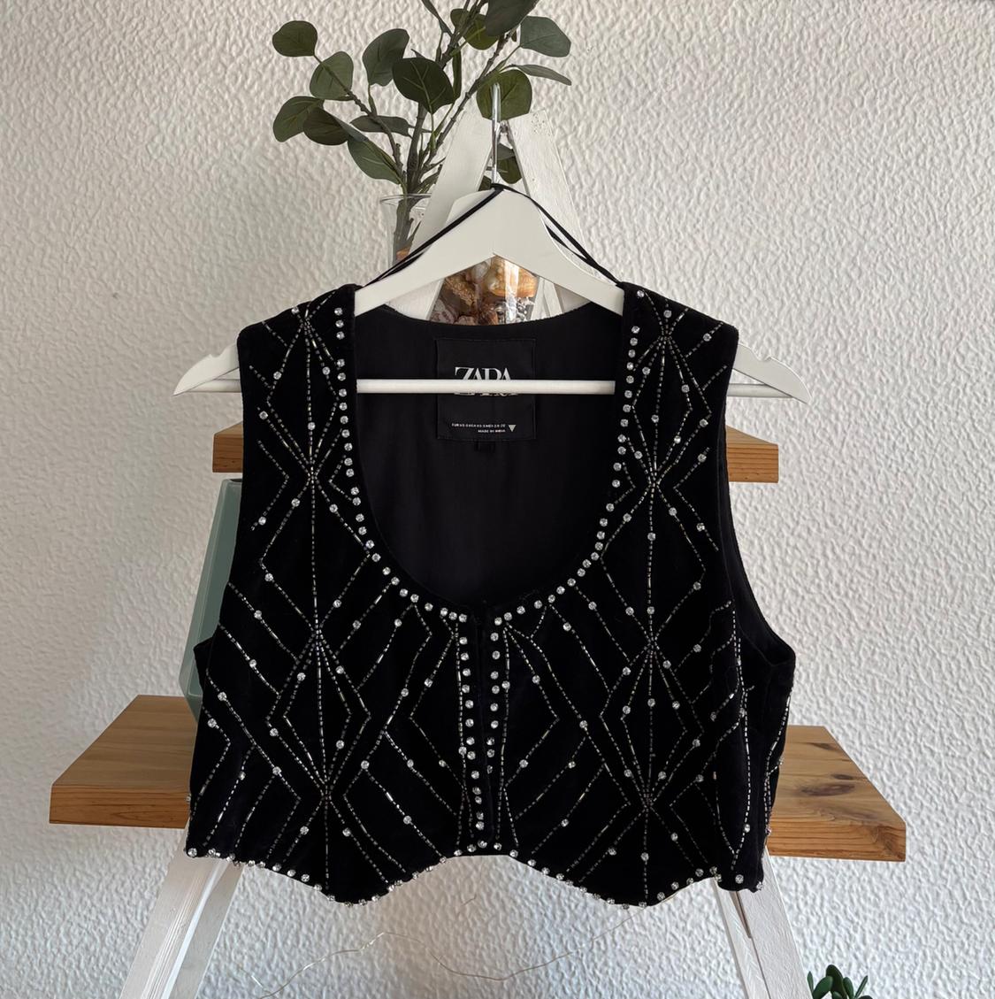 Black waistcoat with pearl details hanging on a white hanger. White wall background and decorative plant. Visible text: "ZARA".