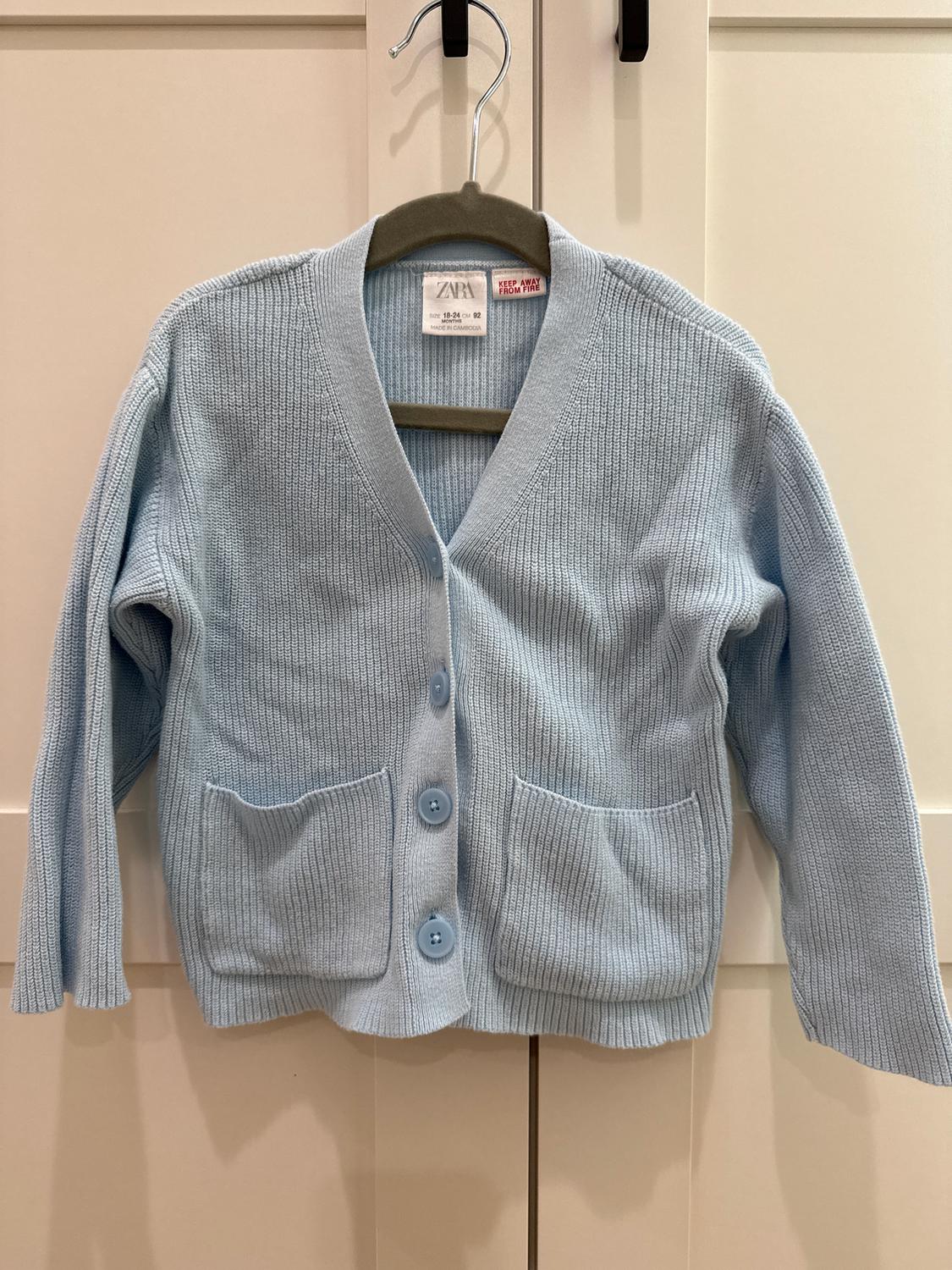 Light blue knitted cardigan with large buttons and two front pockets, hanging on a hanger in front of a white door.