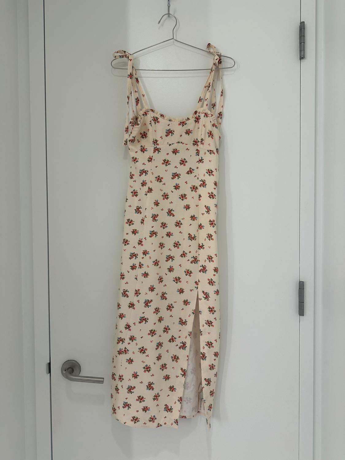 Cream midi dress with floral print, tie straps, and side slit, hanging on a hanger on a white door.