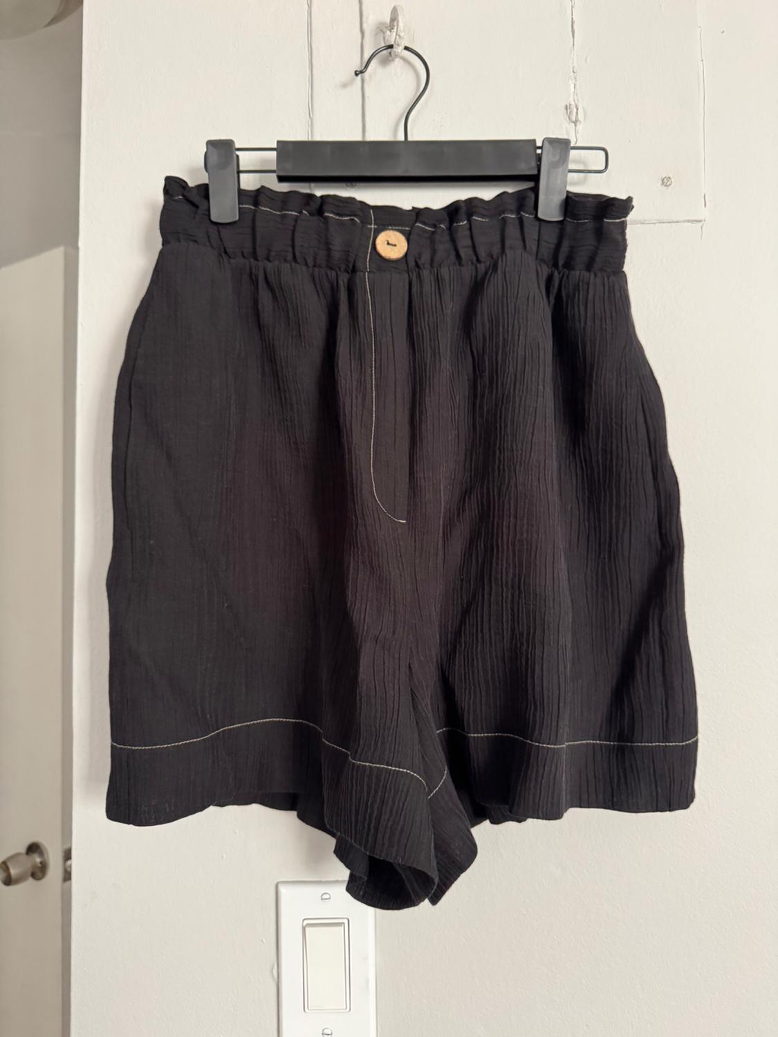 Black shorts with elastic waistband and wooden button, hanging on a black hanger on a white wall.