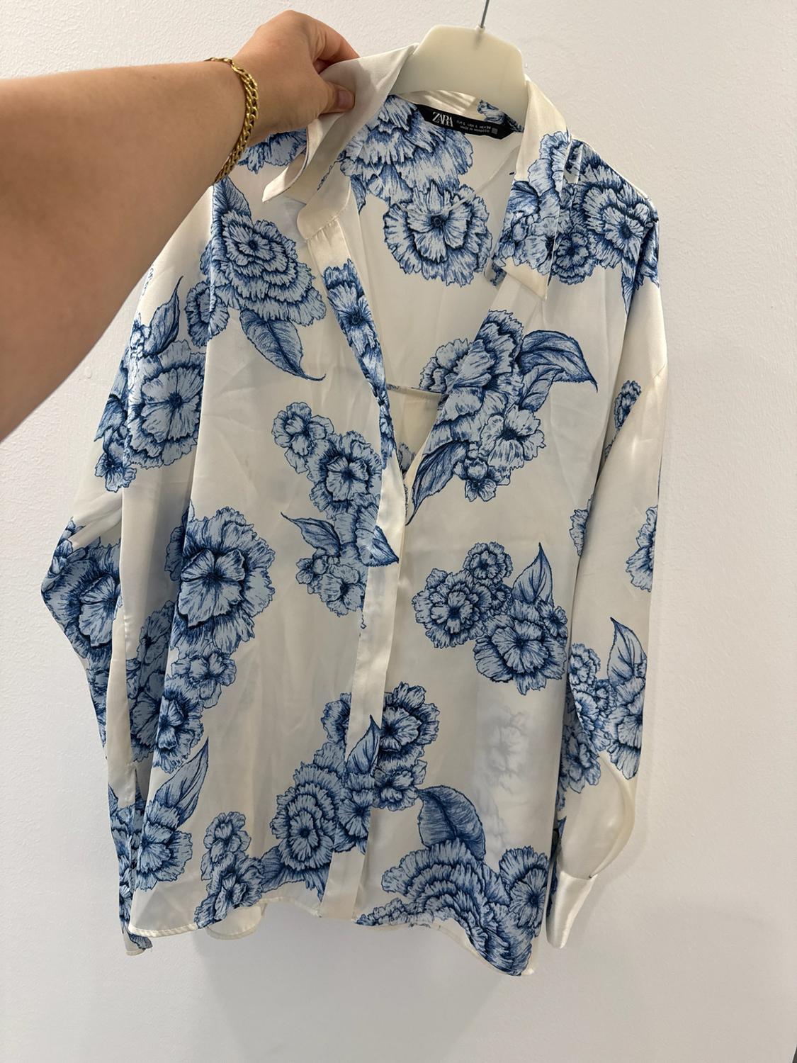 Hand with gold bracelet holding a white shirt with a blue floral print on a hanger in front of a white wall. ZARA label.