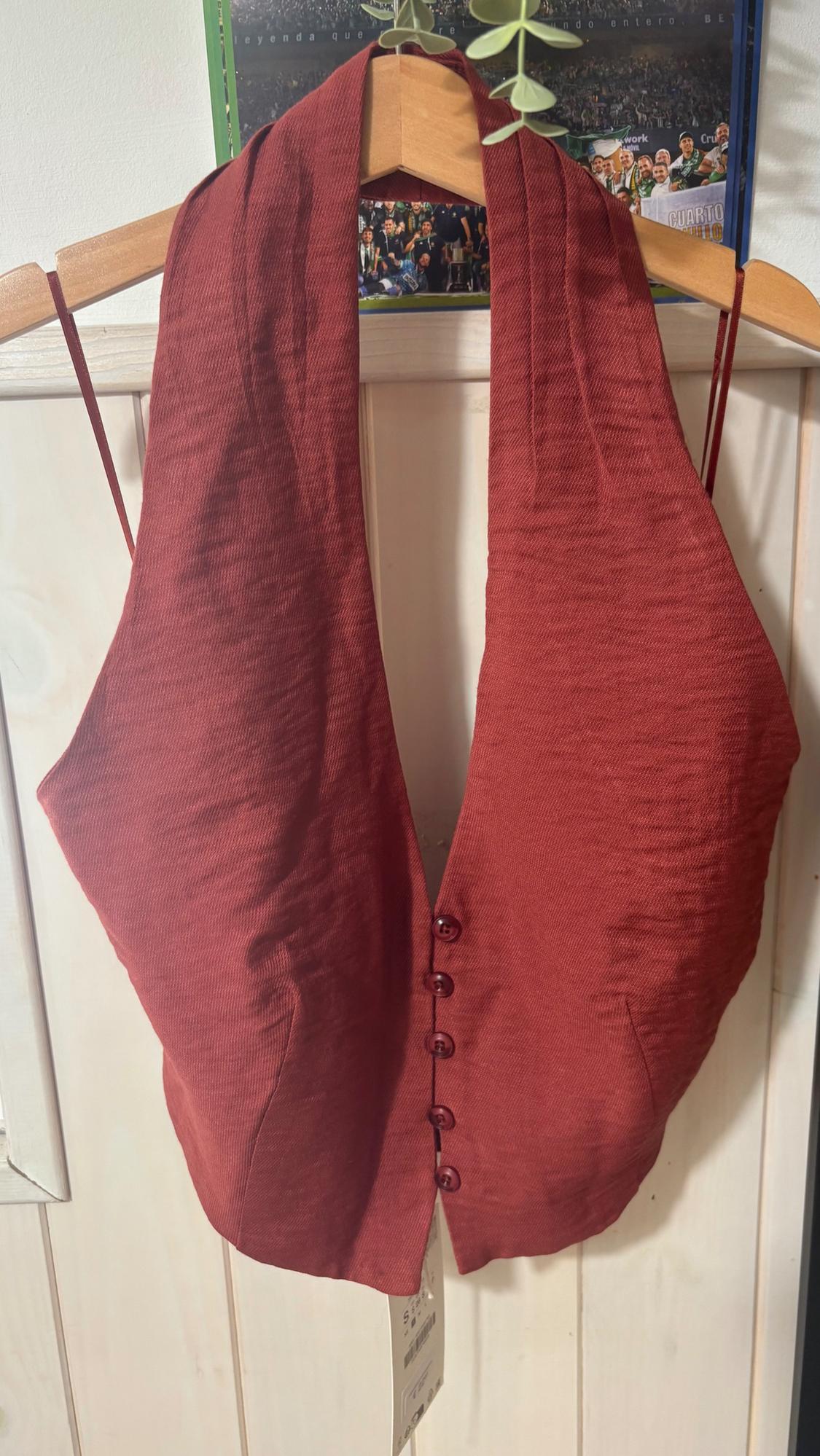 Textured dark red halter-neck waistcoat with front buttons. Hanging on a wooden hanger on a white door, poster in the background.