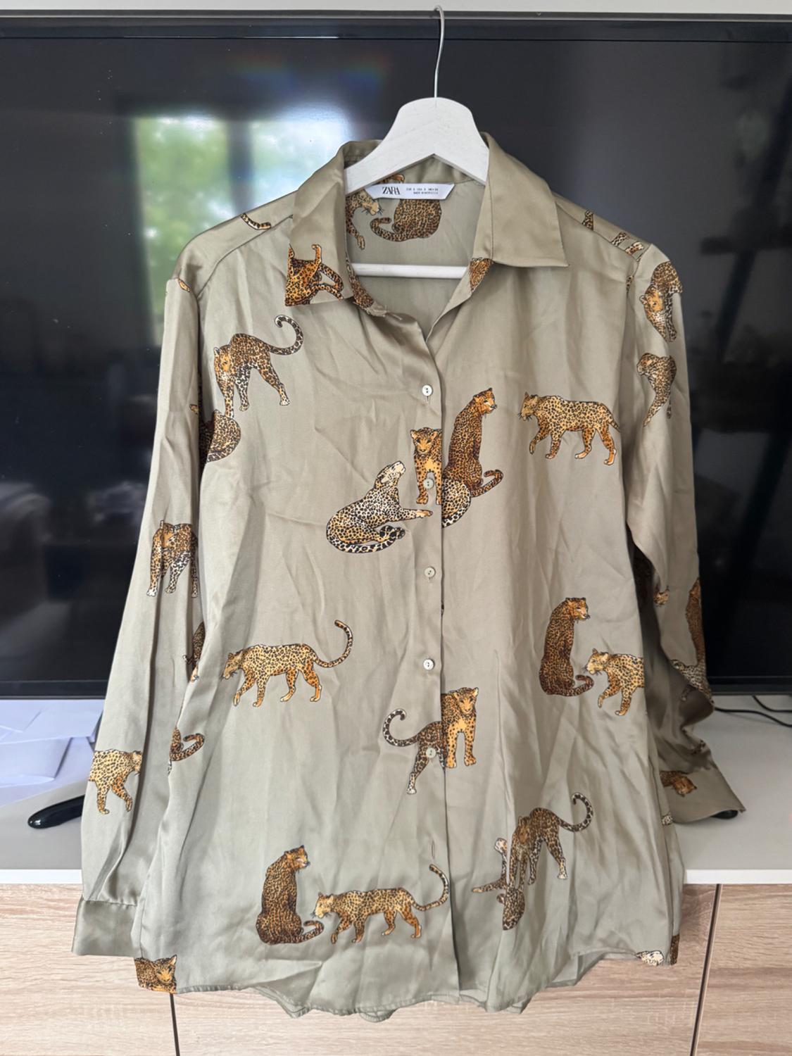 Olive-green long-sleeved shirt with a leopard print, hanging on a white coat hanger in front of a television. Text: ZARA.