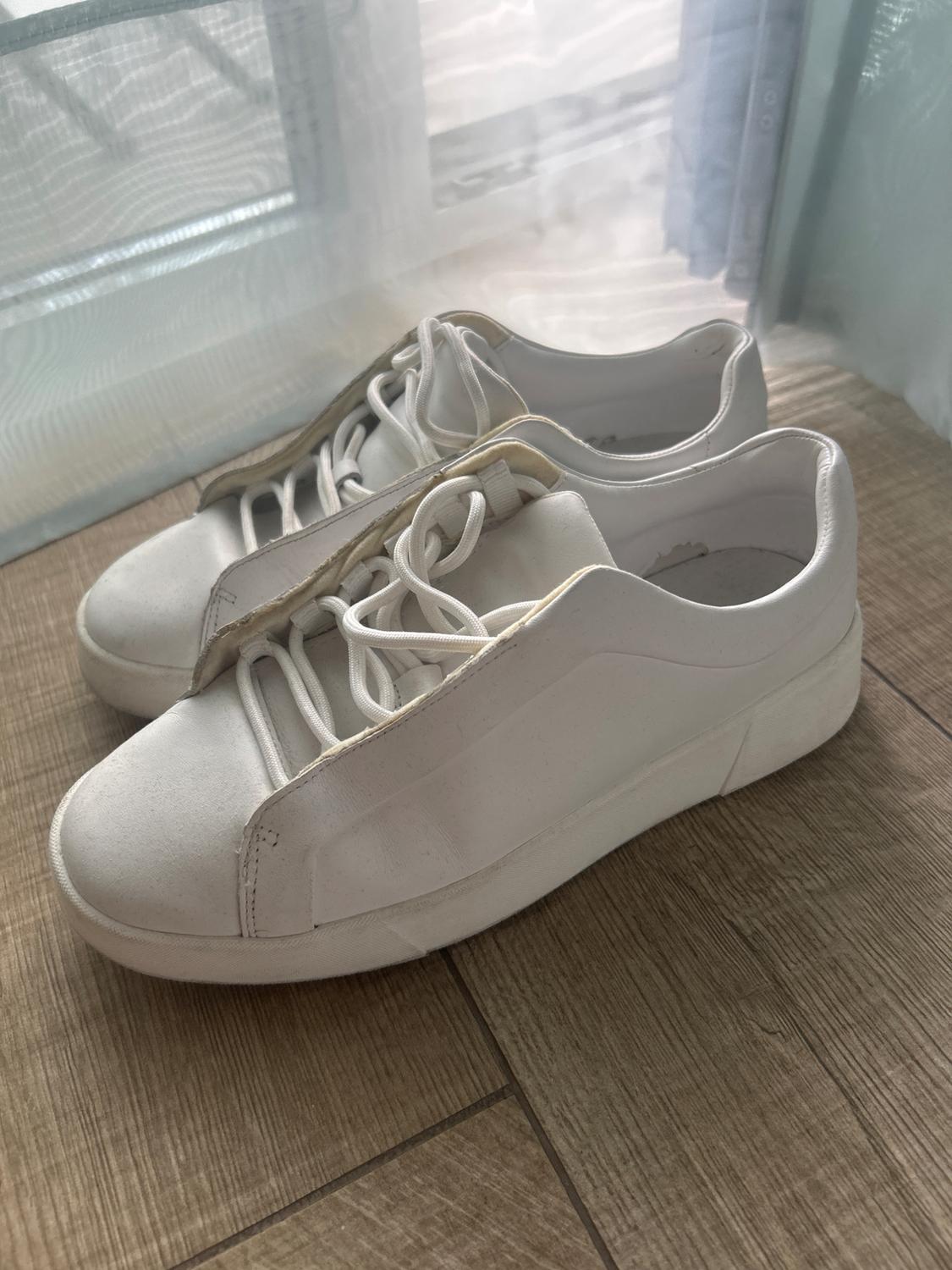White lace-up trainers on a wooden floor next to a window with light-coloured curtains. Casual and relaxed style.