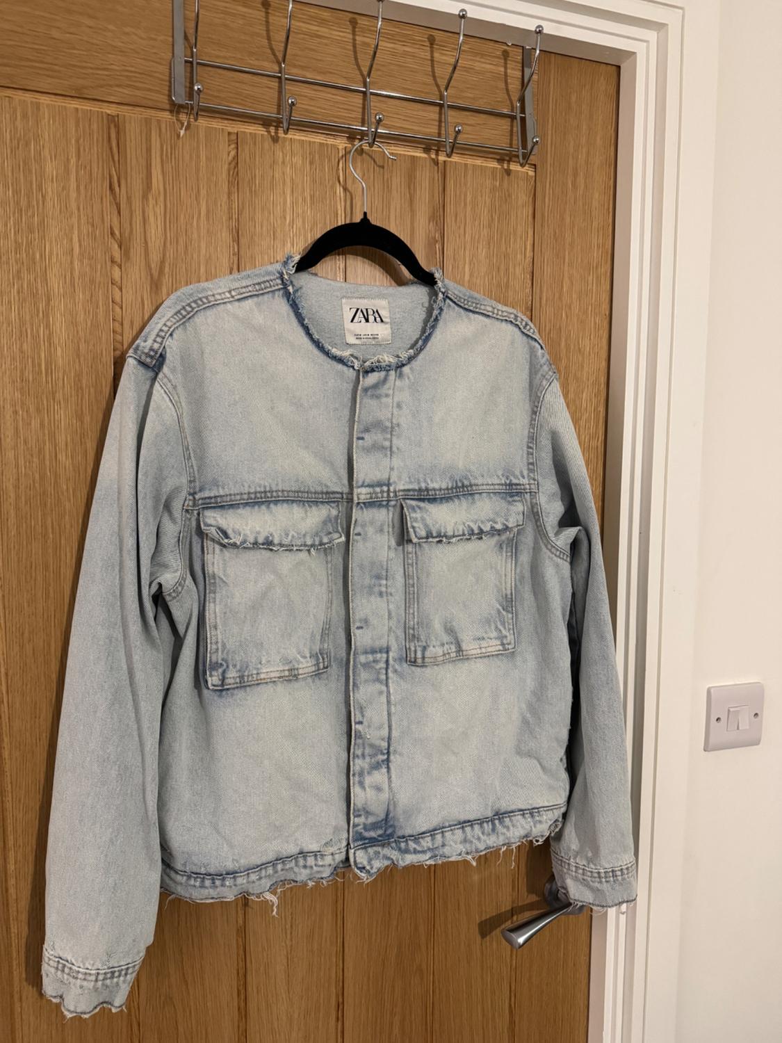 Light blue denim jacket hanging on a wooden door. The label says "ZARA". The jacket has large pockets on the front.
