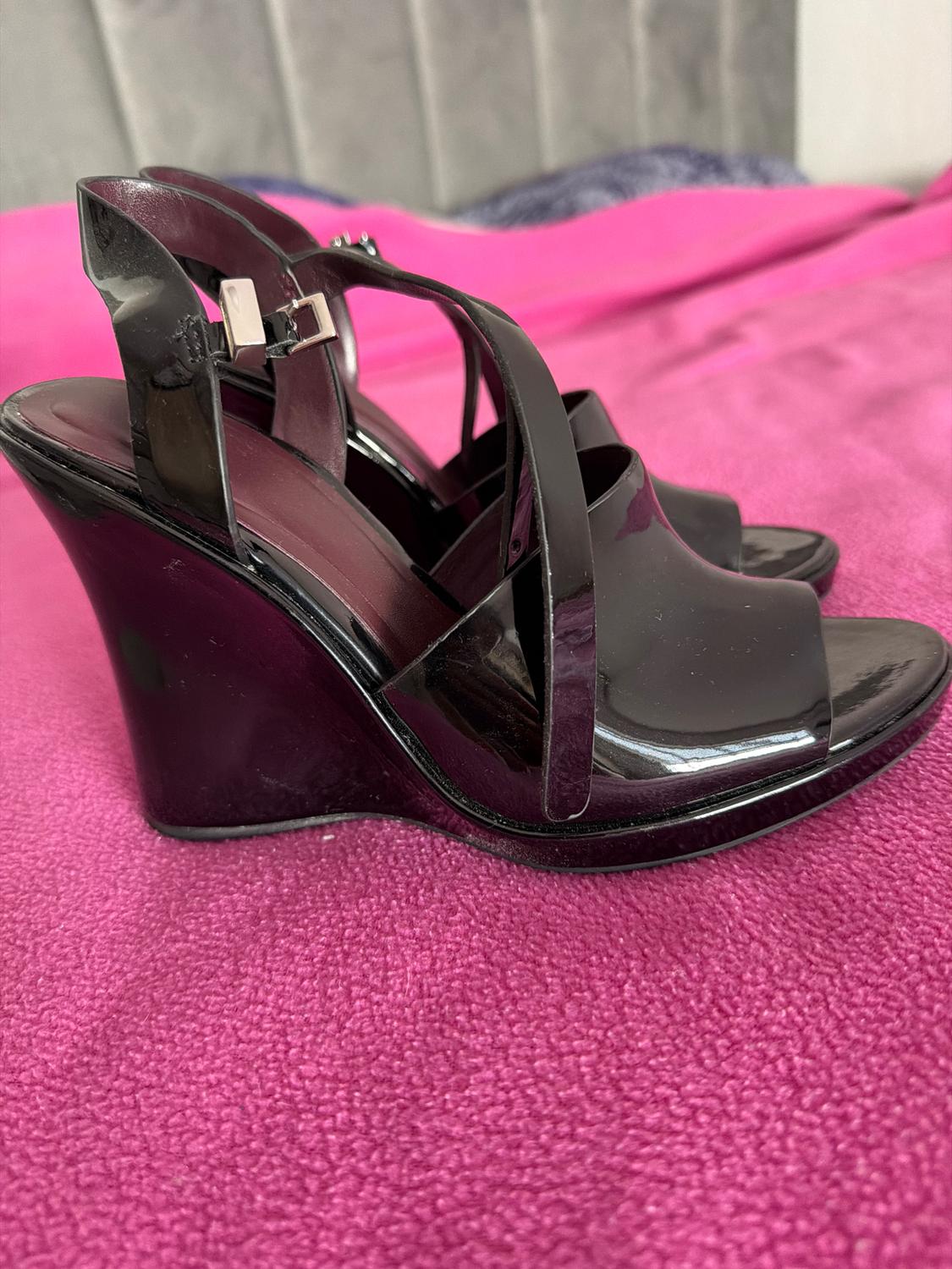 Shiny black buckle-strap high-heeled sandals, on a pink blanket. Grey and blue headboard background.