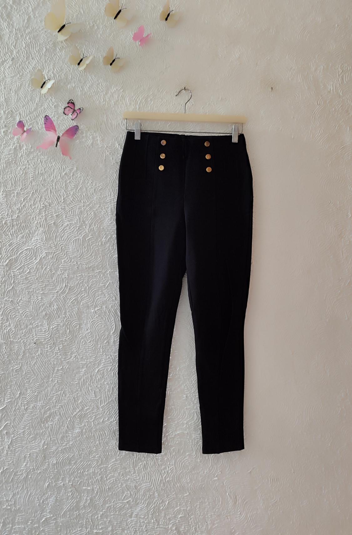 Black trousers with gold buttons hanging on a white textured wall, decorated with paper butterflies in cream and pink tones.
