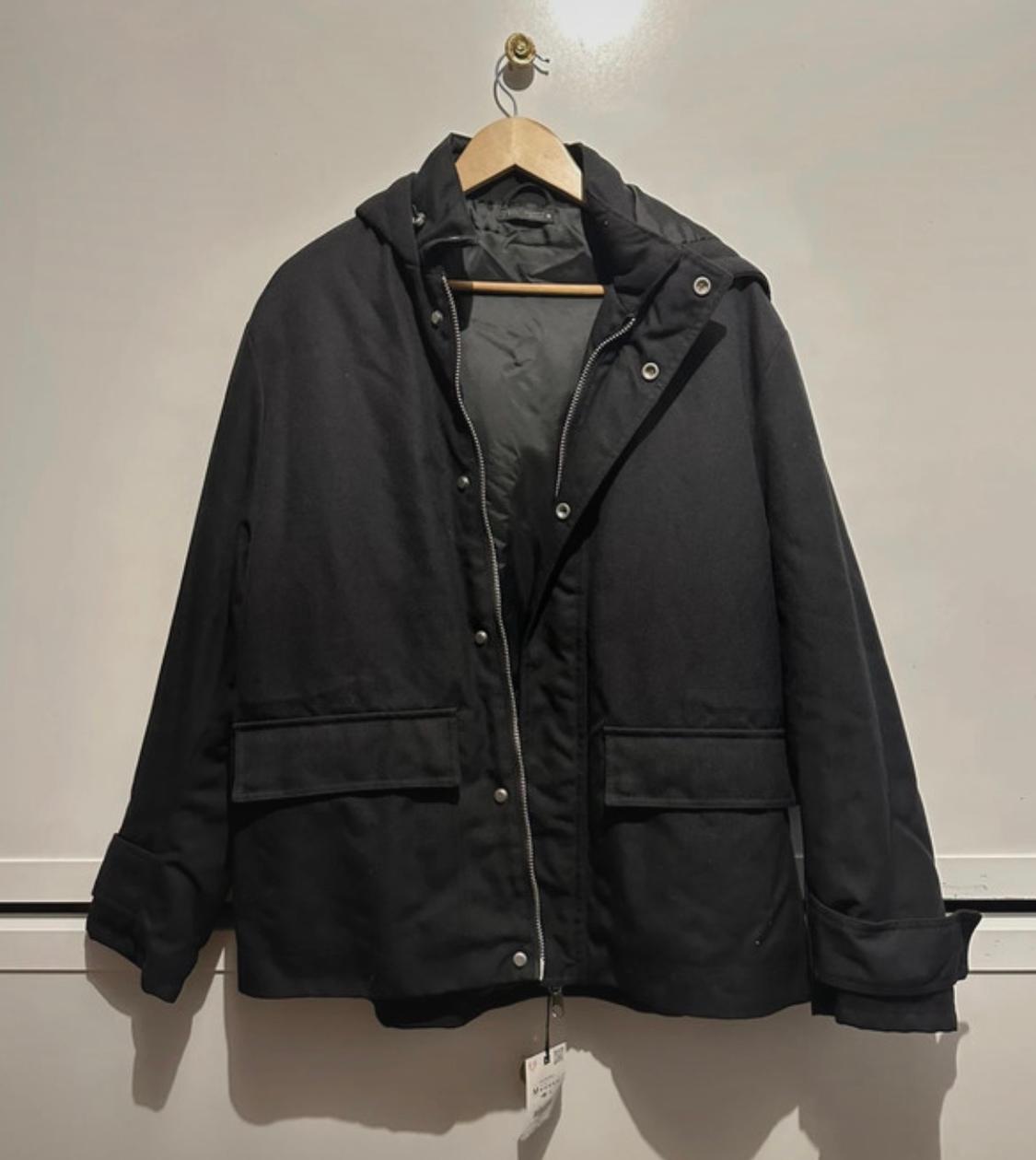 Black hooded jacket hanging on a wooden hanger against a white wall. Label visible at the bottom.