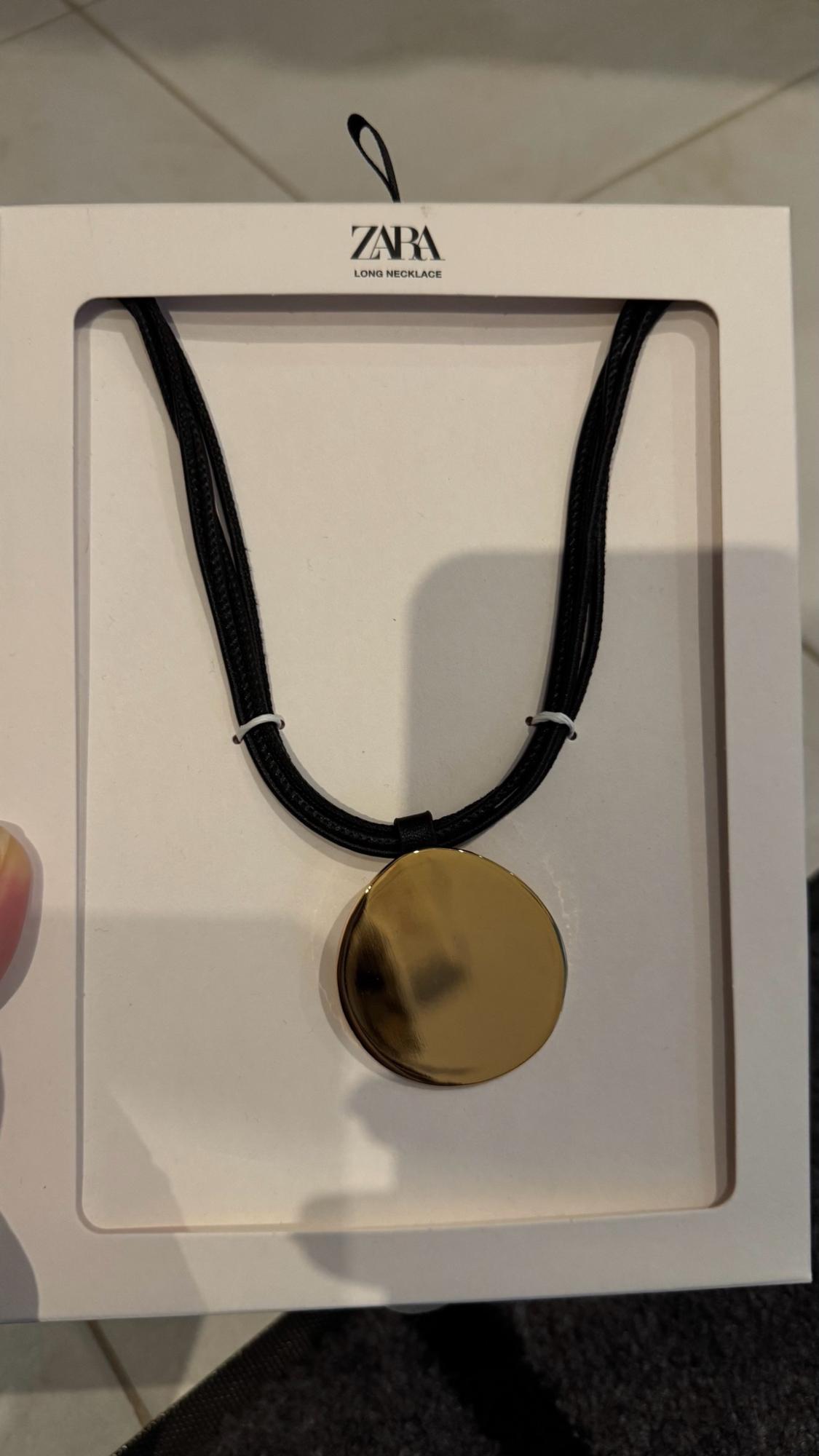 Long black cord necklace with a large gold-tone round pendant in white packaging. Text: ZARA LONG NECKLACE.
