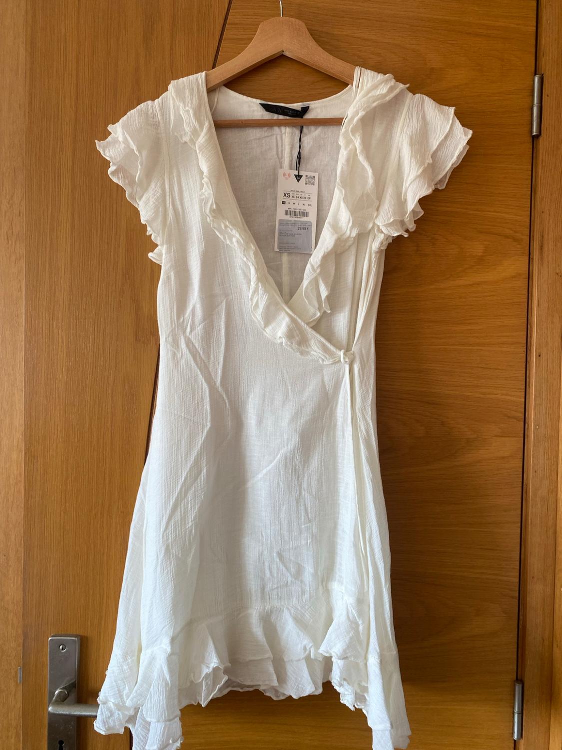 White wrap mini dress with ruffles, hanging from a hanger on a wooden door. Visible label with the text XS.