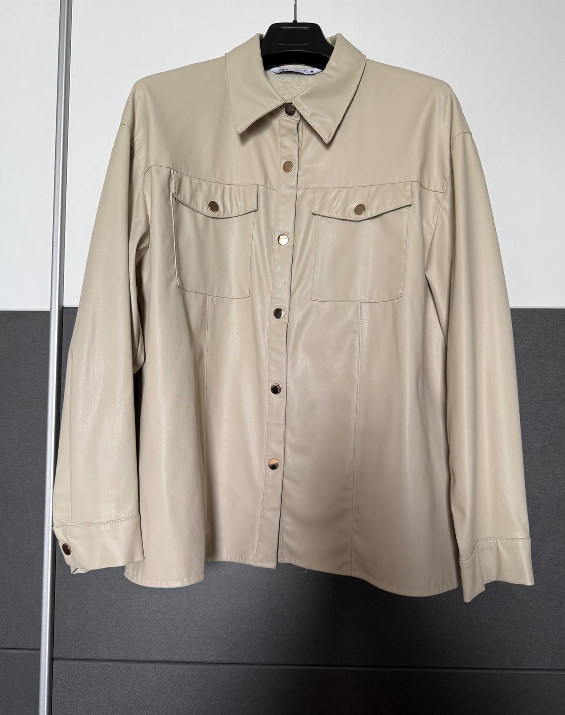 Beige, long-sleeved shirt with metal buttons and chest pockets, hanging on a black hanger against a grey and white background.