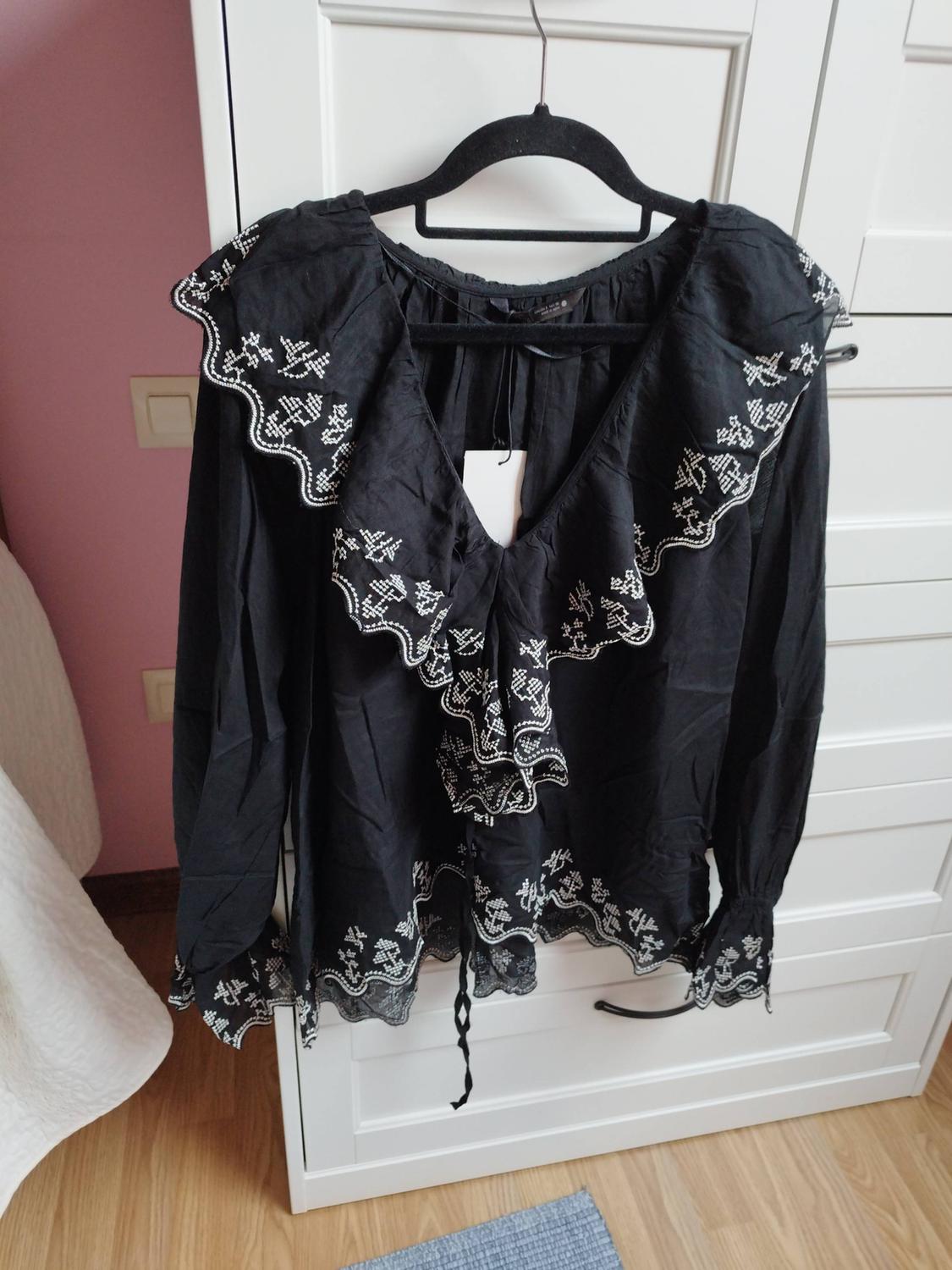 Black blouse with white embroidery, V-neck with ruffles and long sleeves, hanging on a hanger in front of a white wardrobe.