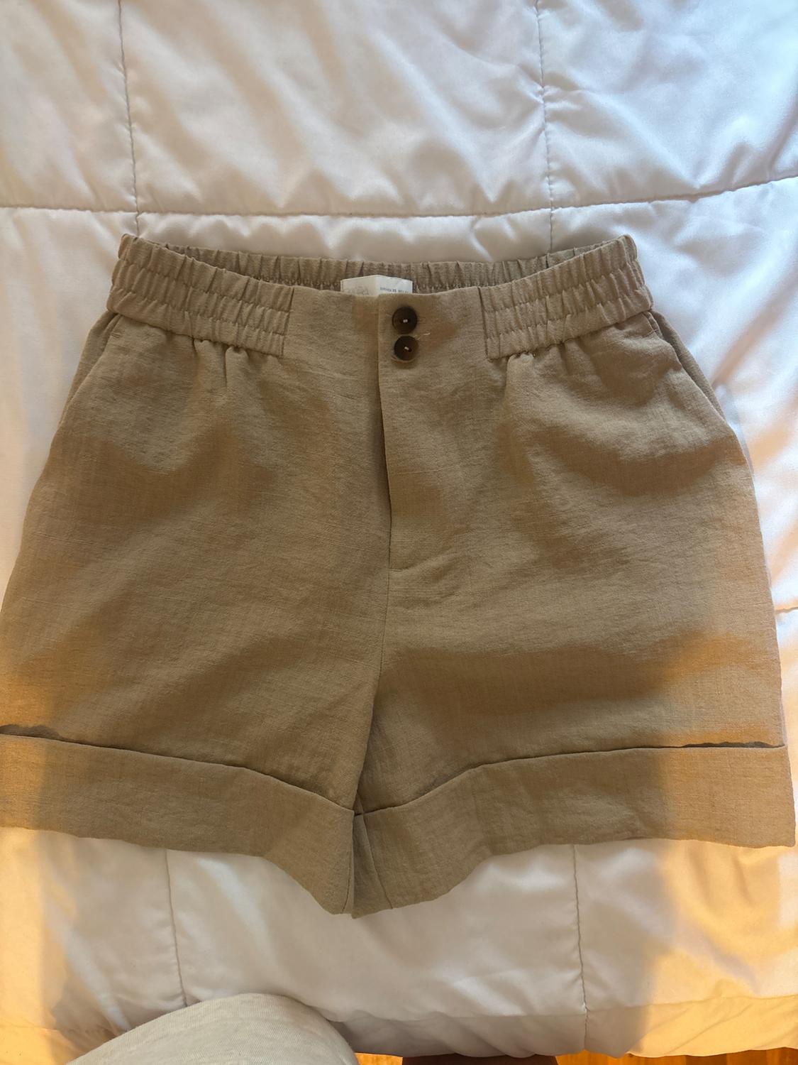 Beige shorts with an elasticated waist, two front buttons, pockets and turn-ups, on a white sheet.