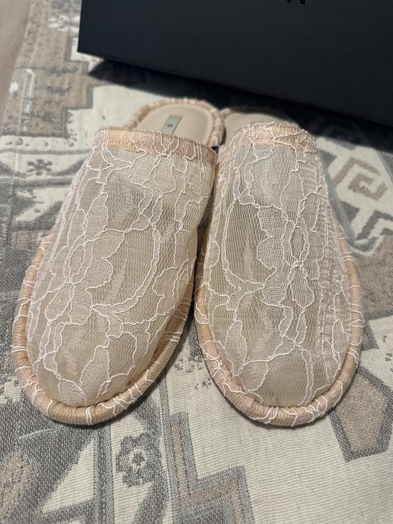 Beige sneakers with white floral lace on a geometric design rug in neutral tones, dark background.