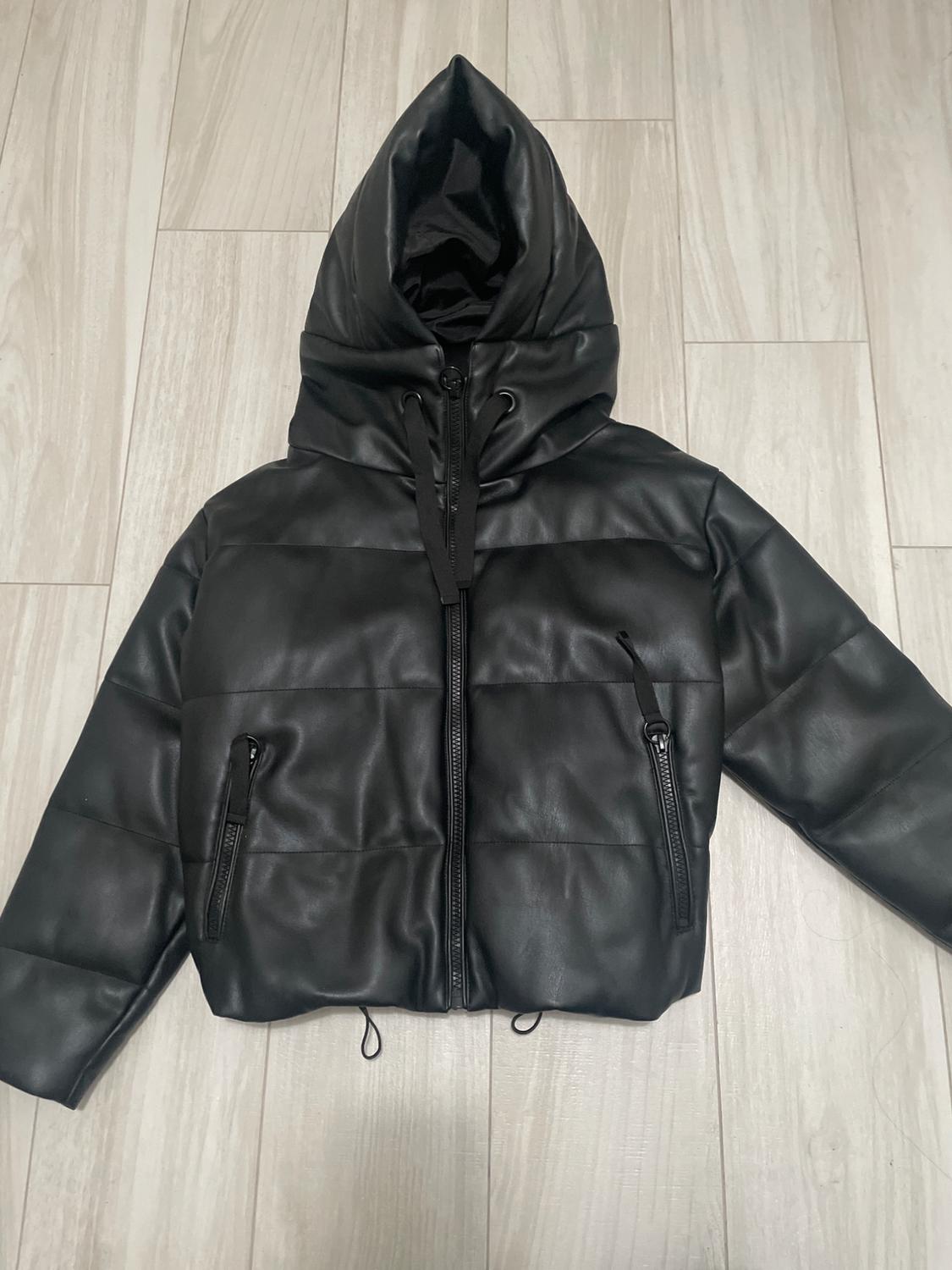Black padded jacket with a hood and front zip, on a light wooden floor. Pockets with visible zips.
