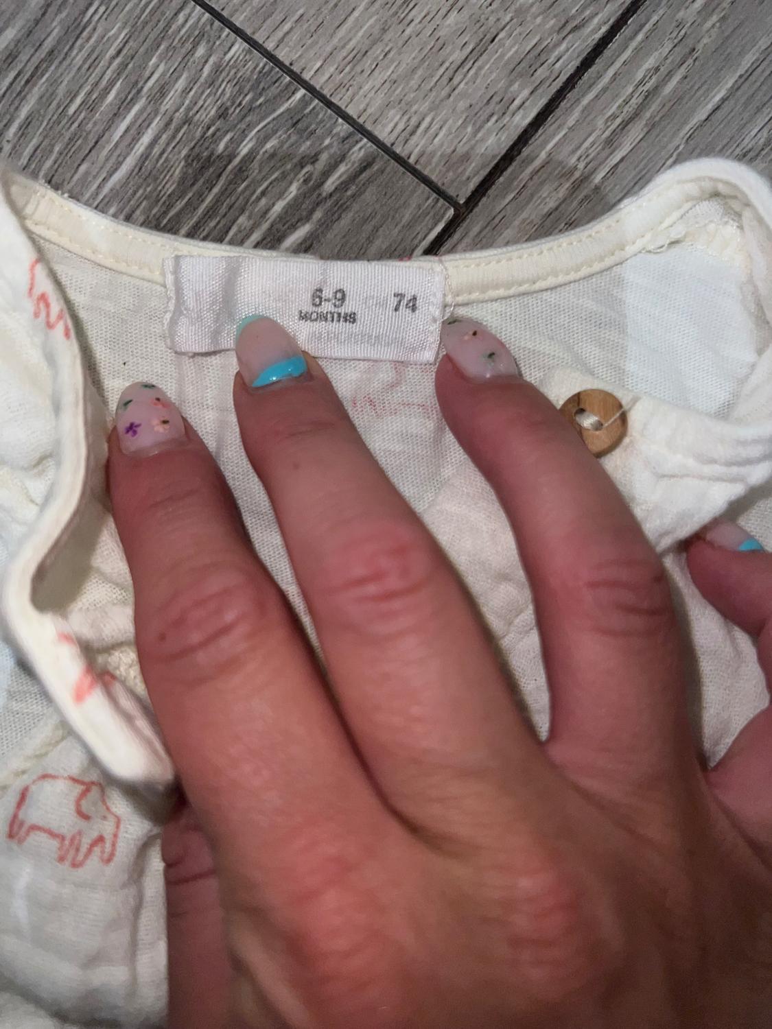 Hand with decorated nails holds a white garment with a label that reads "6-9 MONTHS 74" on a grey wooden floor.