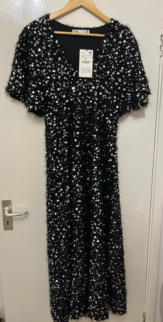 Black dress with silver sequins hanging on a wooden hanger. Visible tag with price of £95. White door in the background.