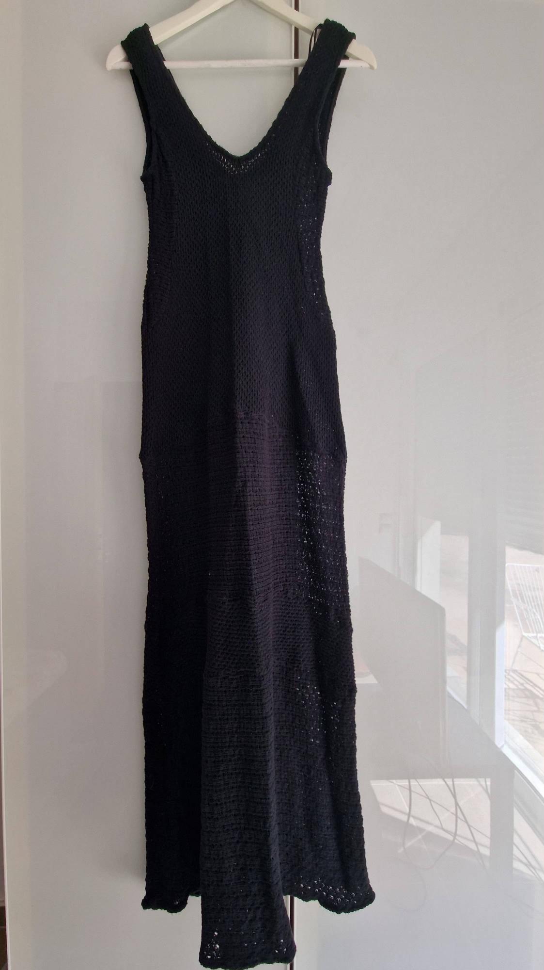 Black sleeveless knitted maxi dress with a V-neck, hanging on a coat hanger in front of a shiny white wardrobe with reflections from a window.
