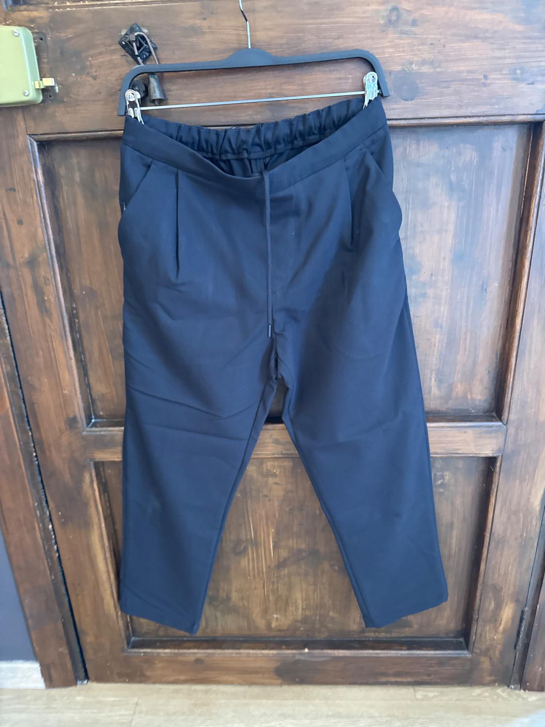 Black trousers hanging on a hanger in front of a wooden door. Simple design with an elasticated waist and front pleats.
