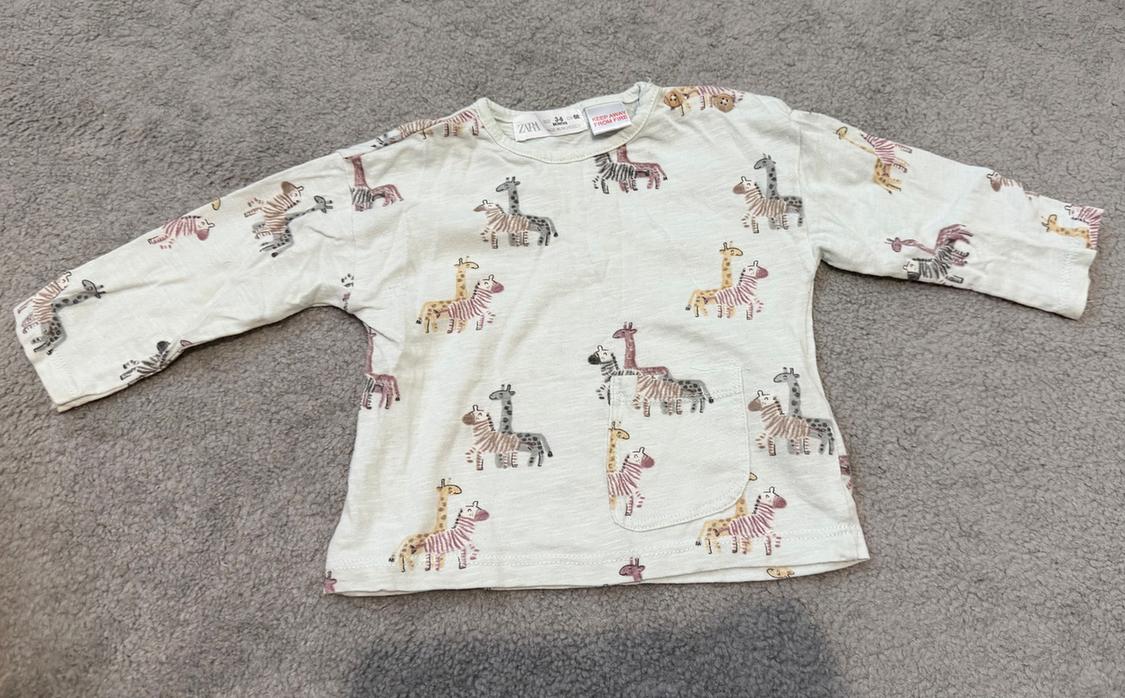 Long-sleeved children's T-shirt with a giraffe and zebra print in pastel shades on a beige background, placed on a grey rug.