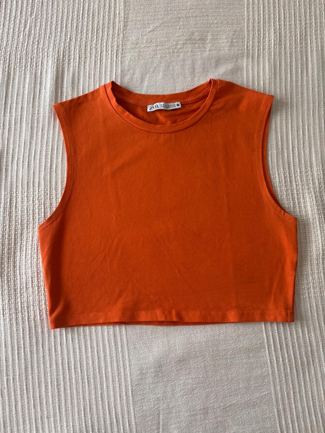 Orange sleeveless crop top on a textured beige surface. Visible label on the collar.