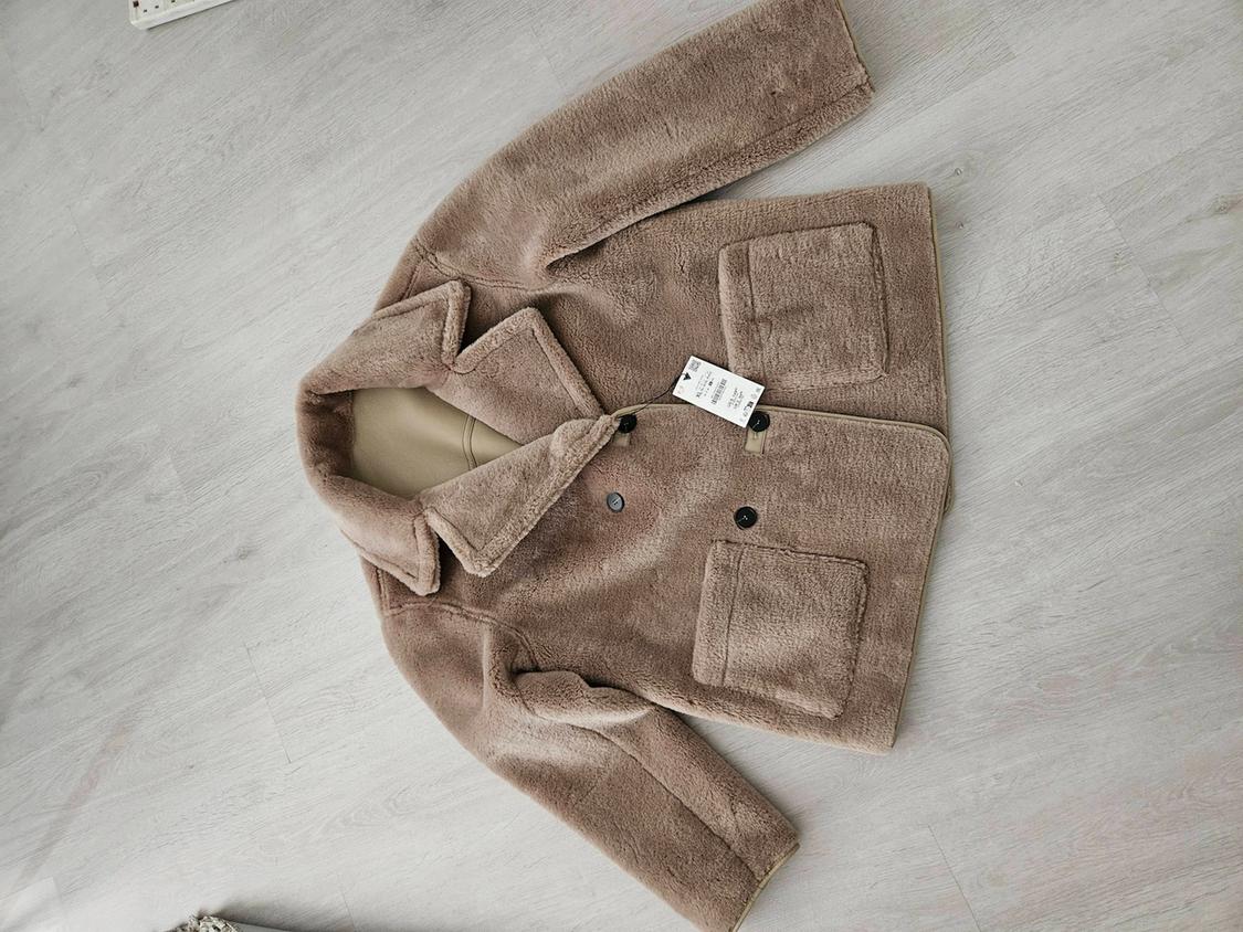 Light brown faux fur jacket with lapels, buttons and pockets, laid out in a relaxed manner on a wooden floor.