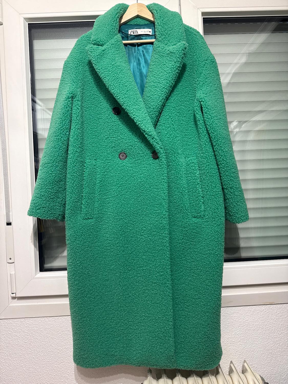 Green wool coat with black buttons hanging on a hanger in front of a window with white blinds. Visible label.