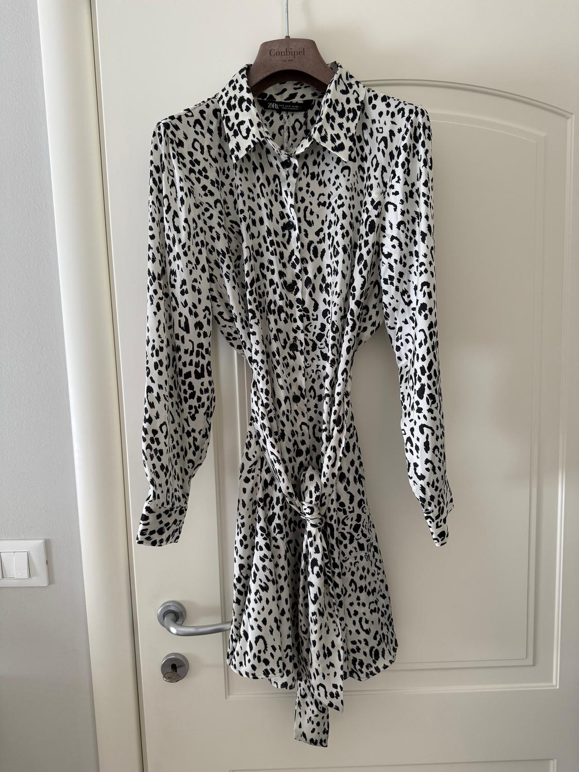 Black and white leopard print long-sleeved dress hanging on a white door. Wooden hanger with "Conbipel" text.