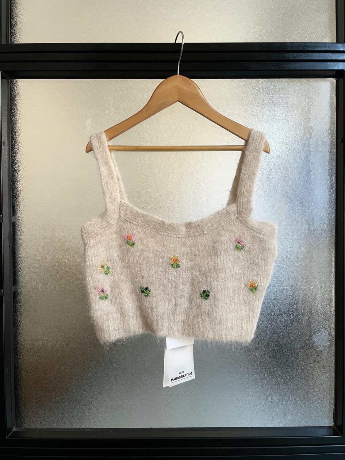Μπεζ πλεκτό crop top με μικρά πολύχρωμα λουλούδια, κρεμασμένο σε ξύλινη κρεμάστρα μπροστά από ημιδιαφανές κρύσταλλο.