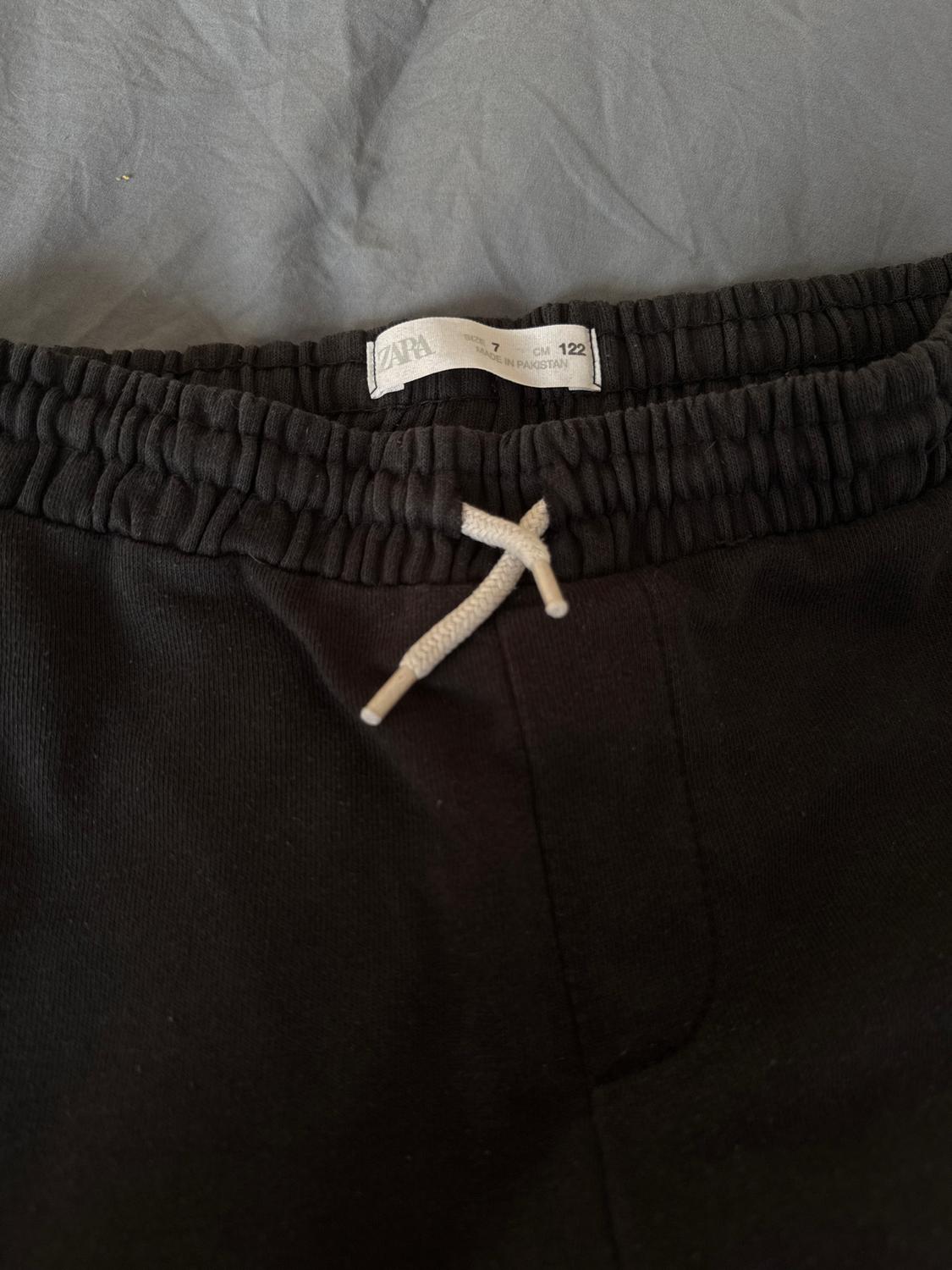 Black trousers with an elasticated waist and white drawstring on a grey background. Visible label: "ZARA, Size 7, 122 CM, Made in Pakistan".