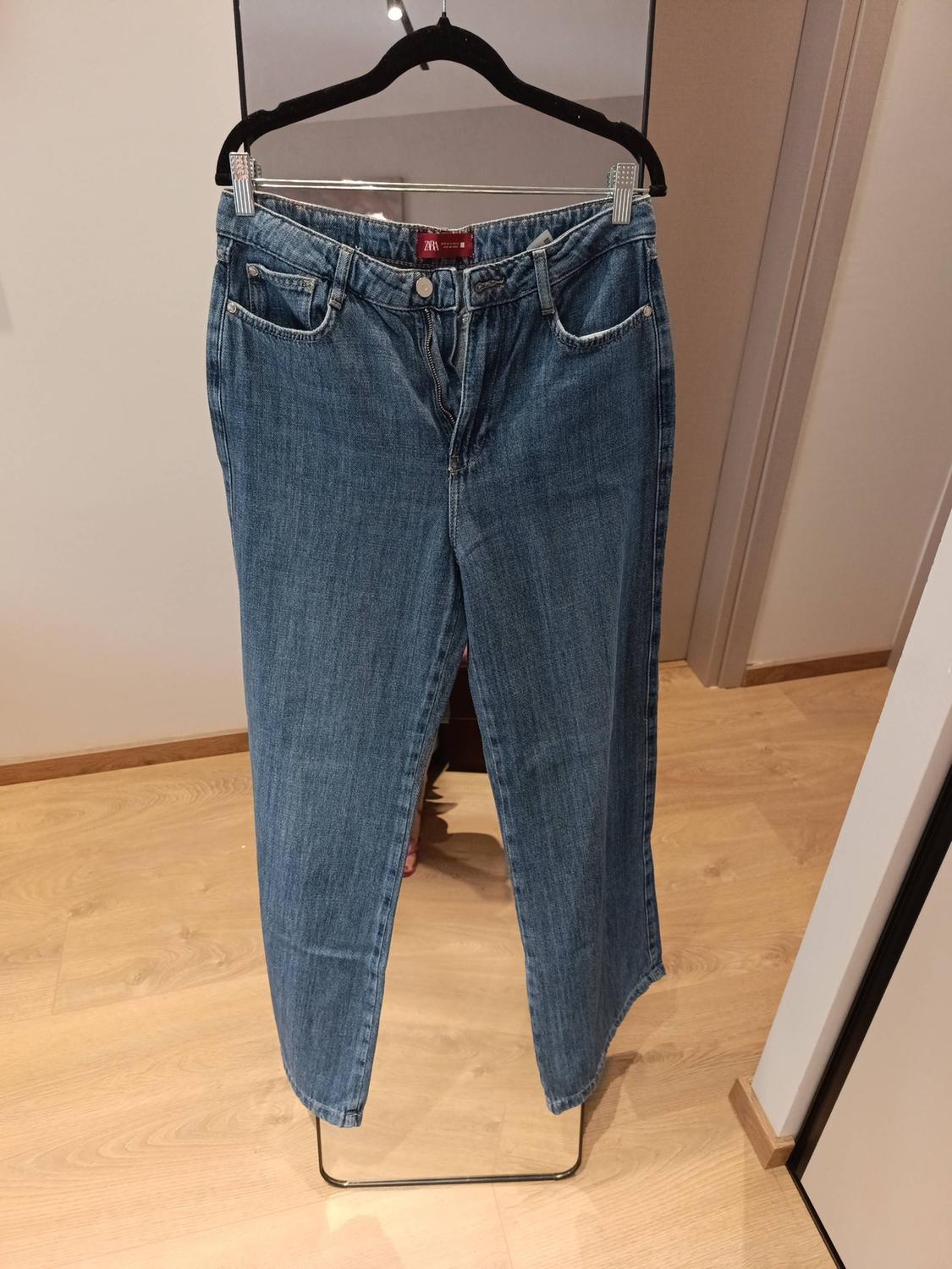 Blue jeans hanging on a black hanger in a room with a wooden floor. The inside label shows the text ZARA.