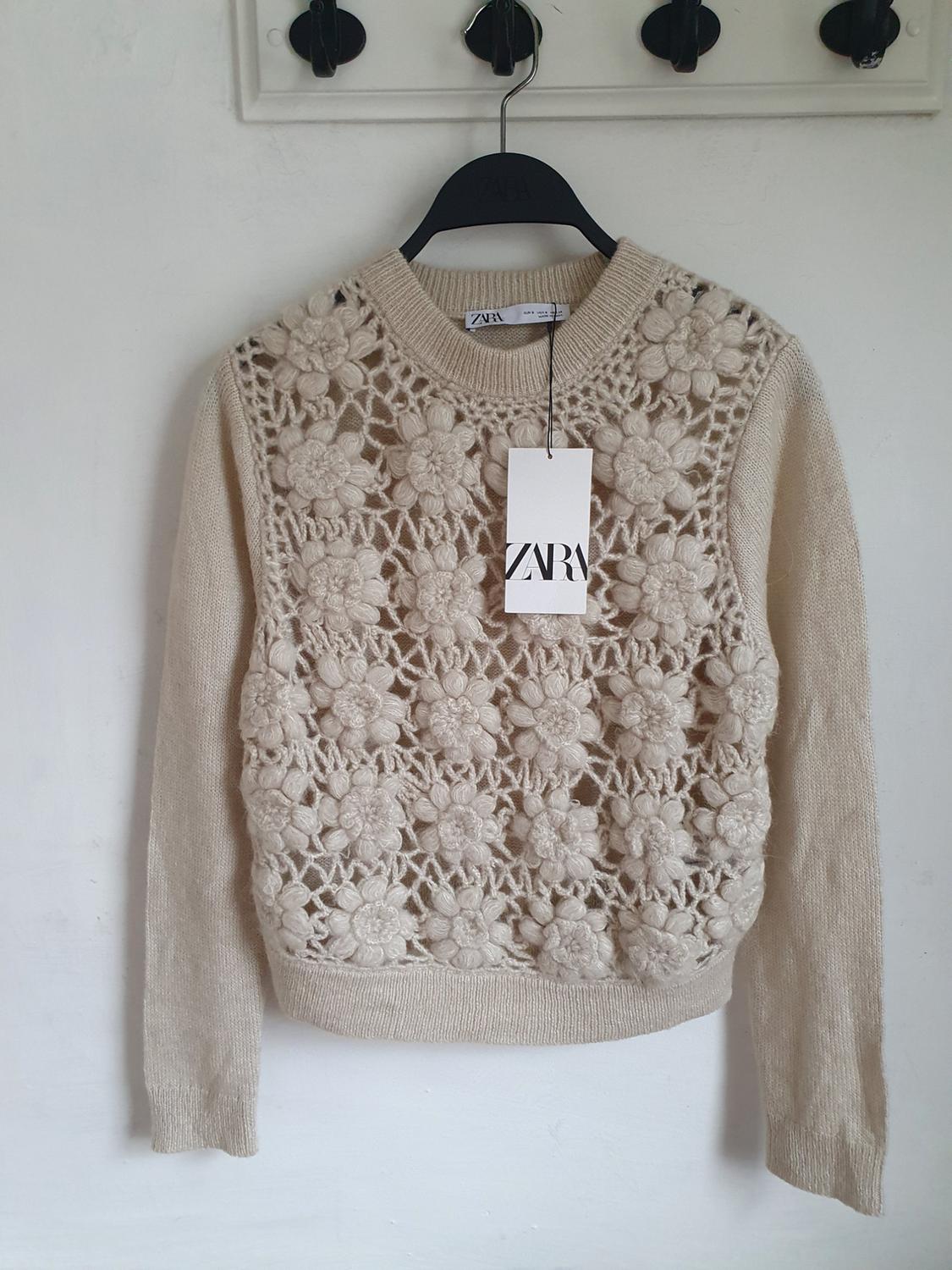 Beige jumper with an openwork floral design, hanging on a black hanger with a Zara label. White background and hooks on the wall.