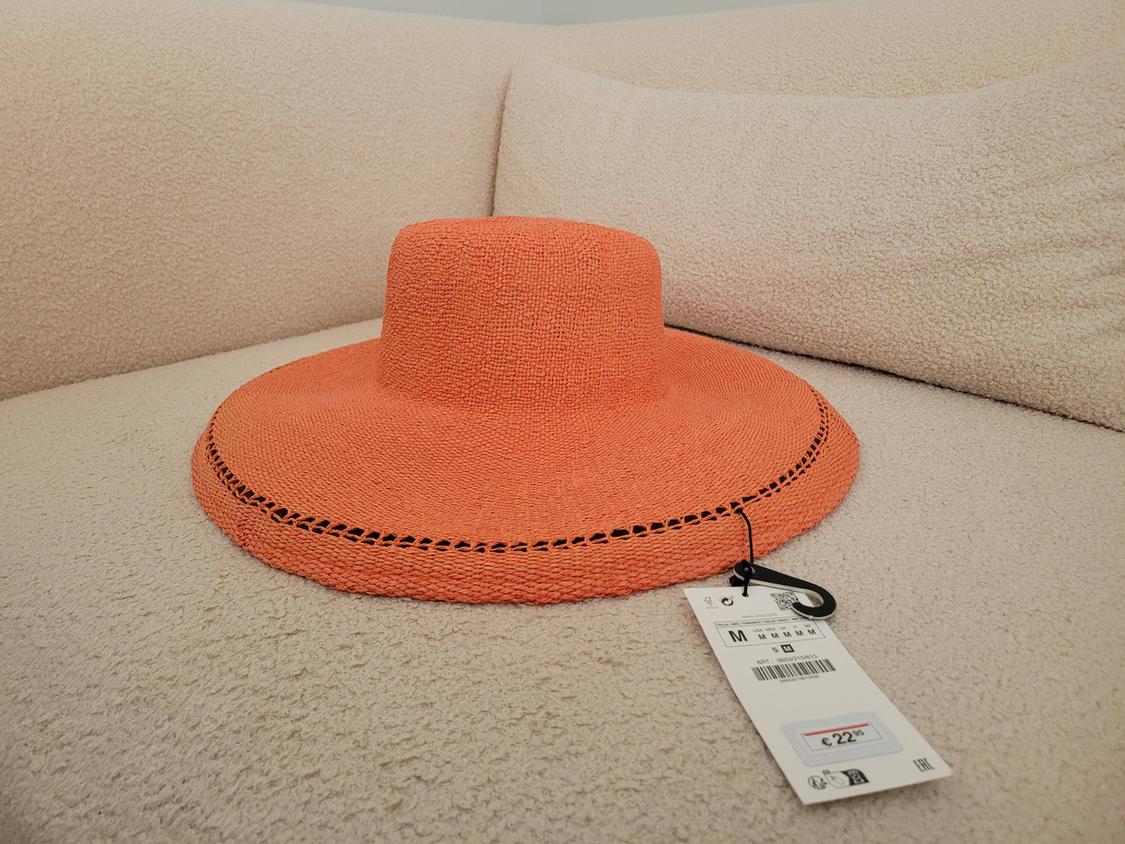Wide-brimmed orange hat on a beige sofa. Visible tag with price of €22.95. Relaxed and modern atmosphere.