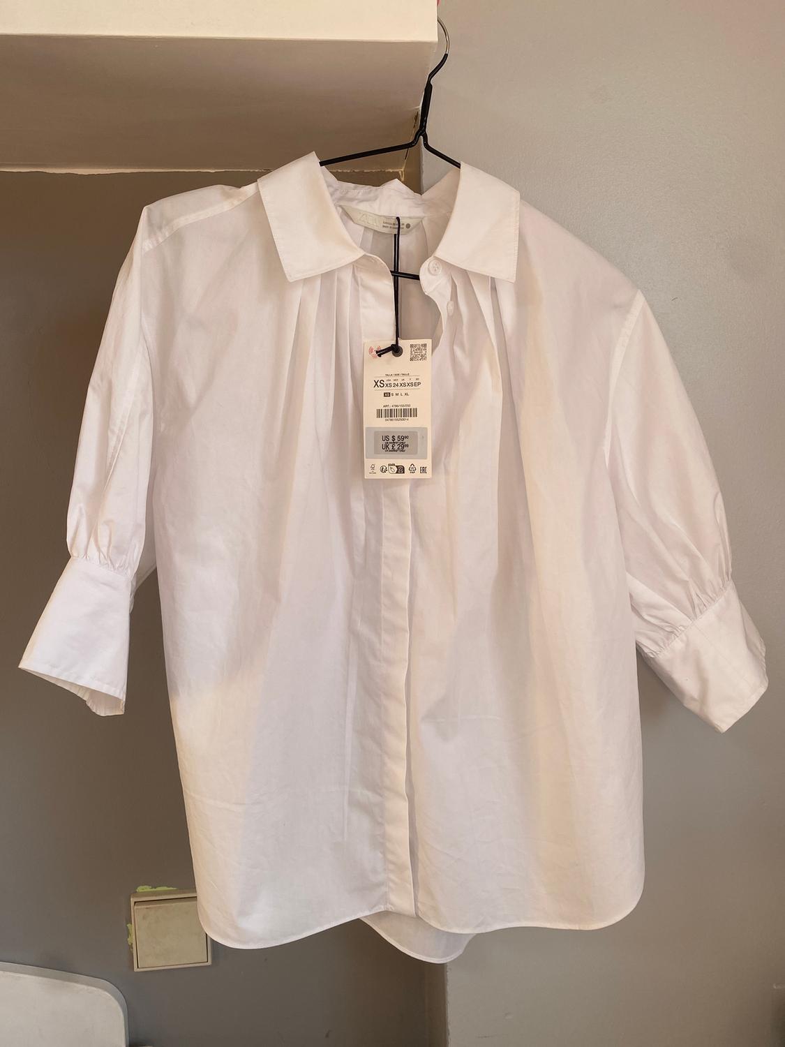 White long-sleeved shirt hanging on a black hanger. Visible label with XS size details and price in dollars. Beige background.
