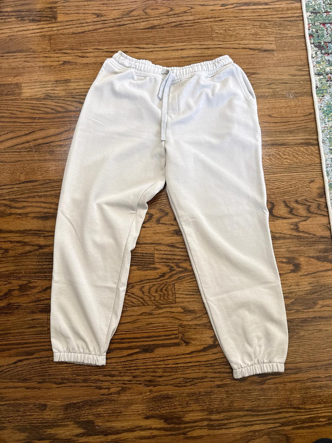 Light beige track pants with an elastic waistband, an adjustable drawstring, and cuffs at the ankles, on a wooden floor.
