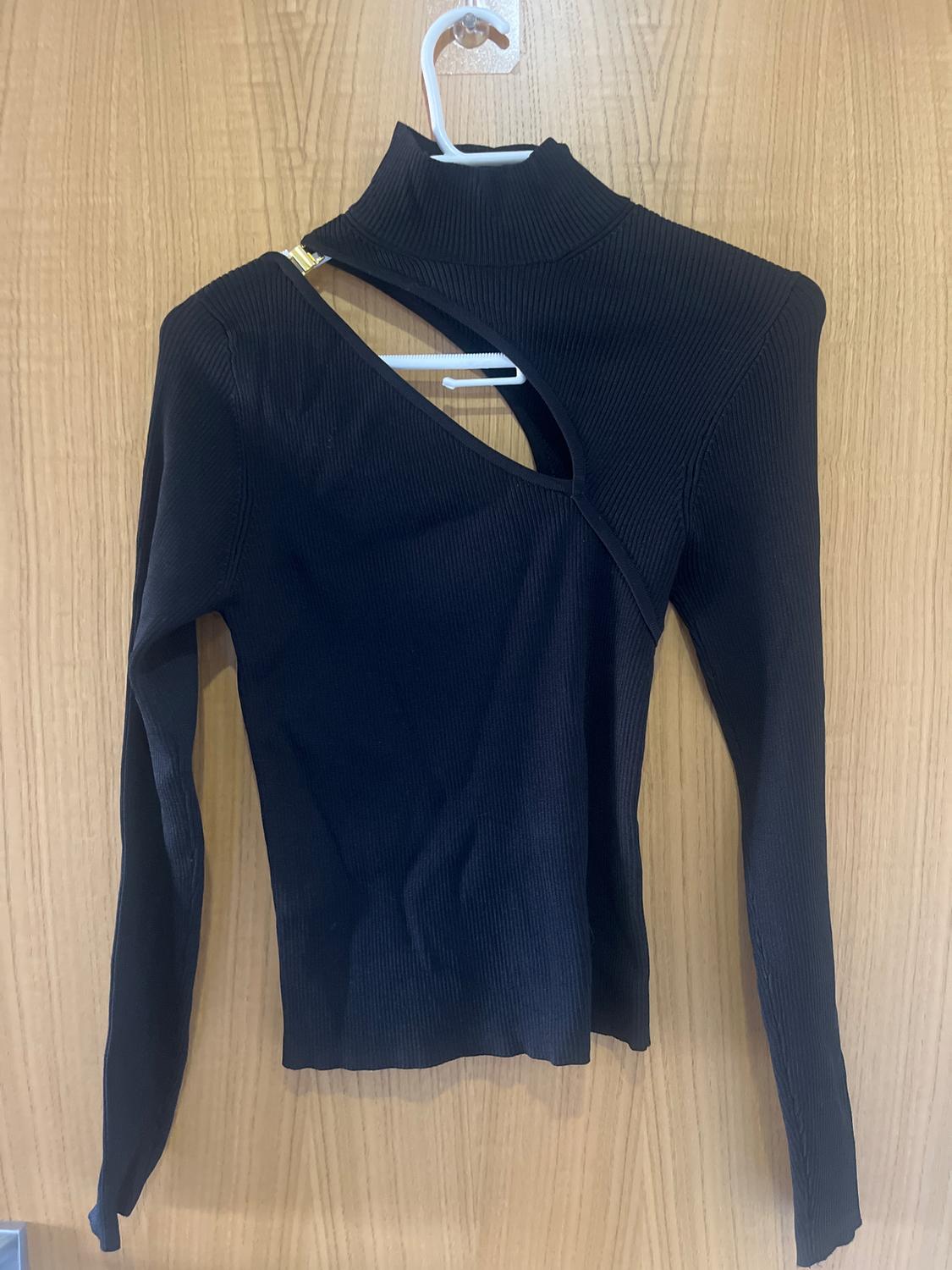 Black long-sleeved polo neck top with an asymmetric design, hanging on a white hanger over a wooden door.