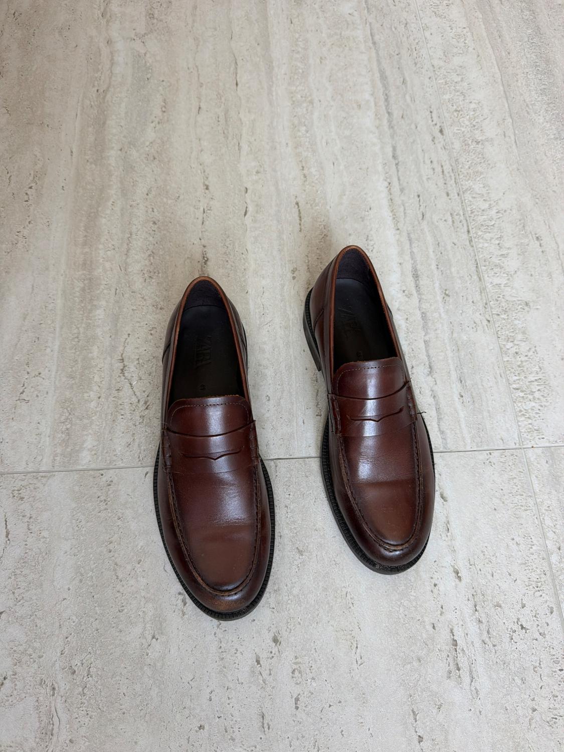 Top view of a pair of brown leather loafers on a light stone floor. Inside, it reads ZARA.