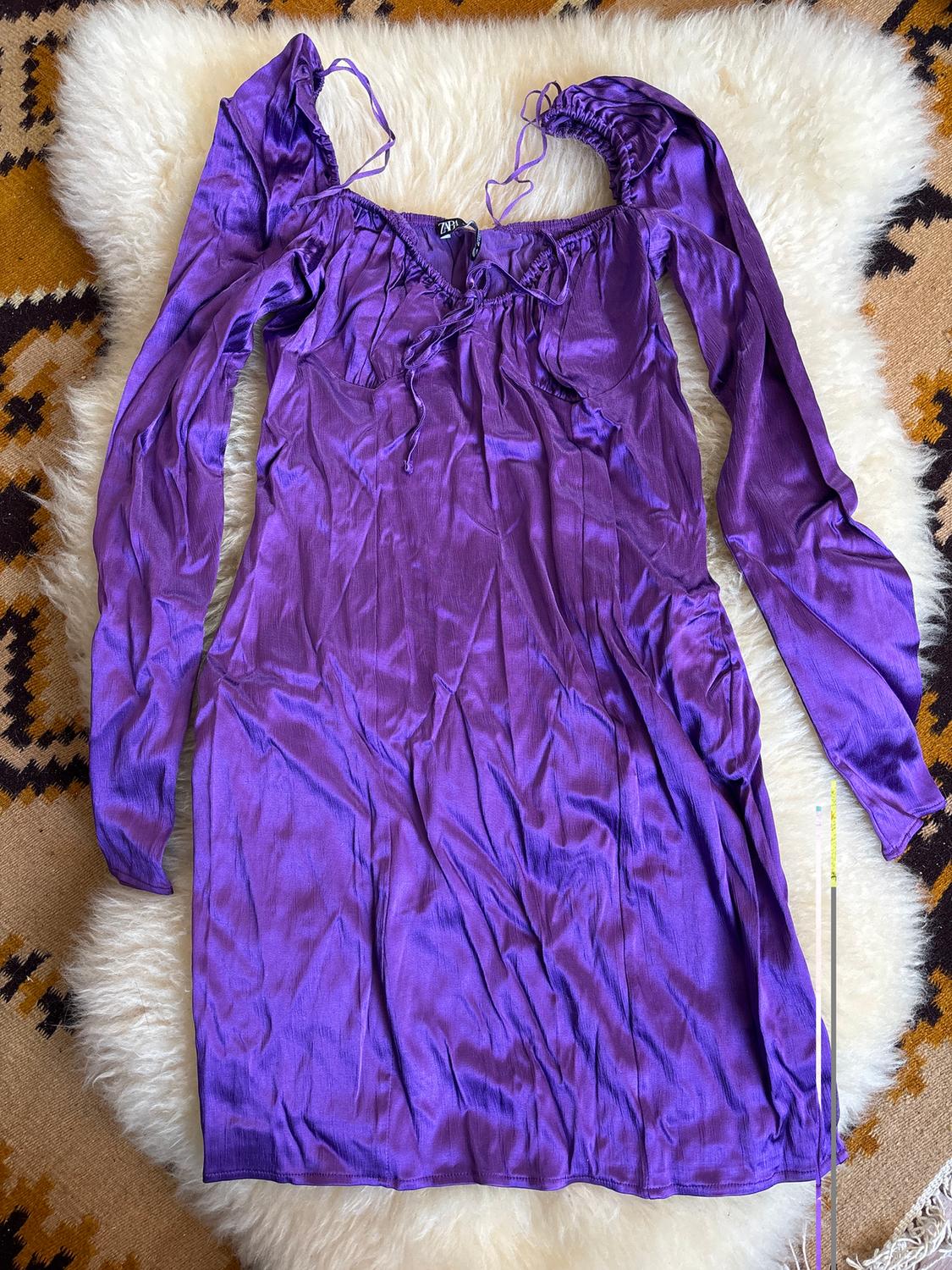 Purple long-sleeved dress with ruched neckline, on a white rug and a background of a rug with a brown and black geometric pattern.