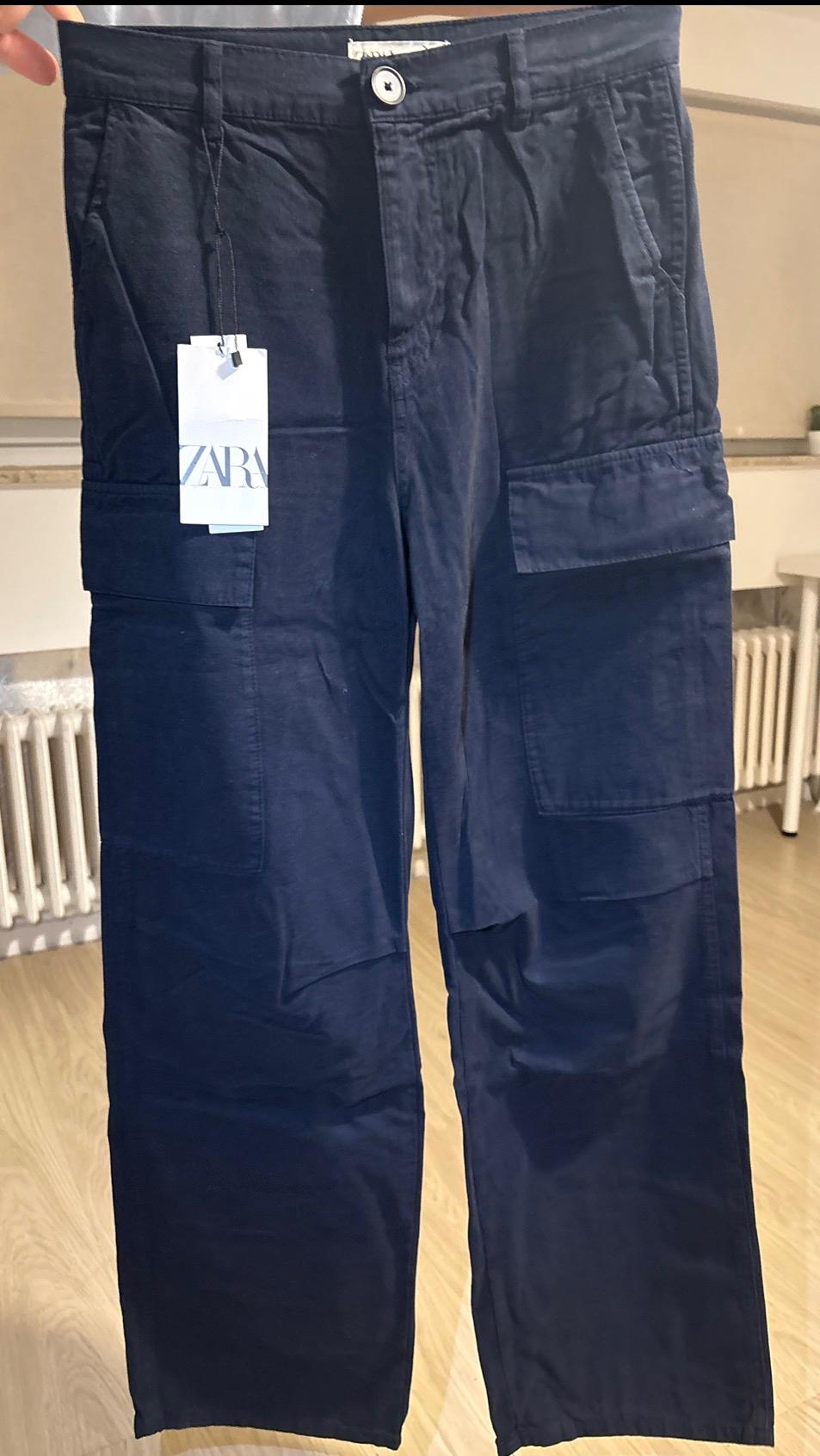 Dark blue cargo trousers, hung up, with large pockets and a visible Zara label. Interior background with light wooden flooring.