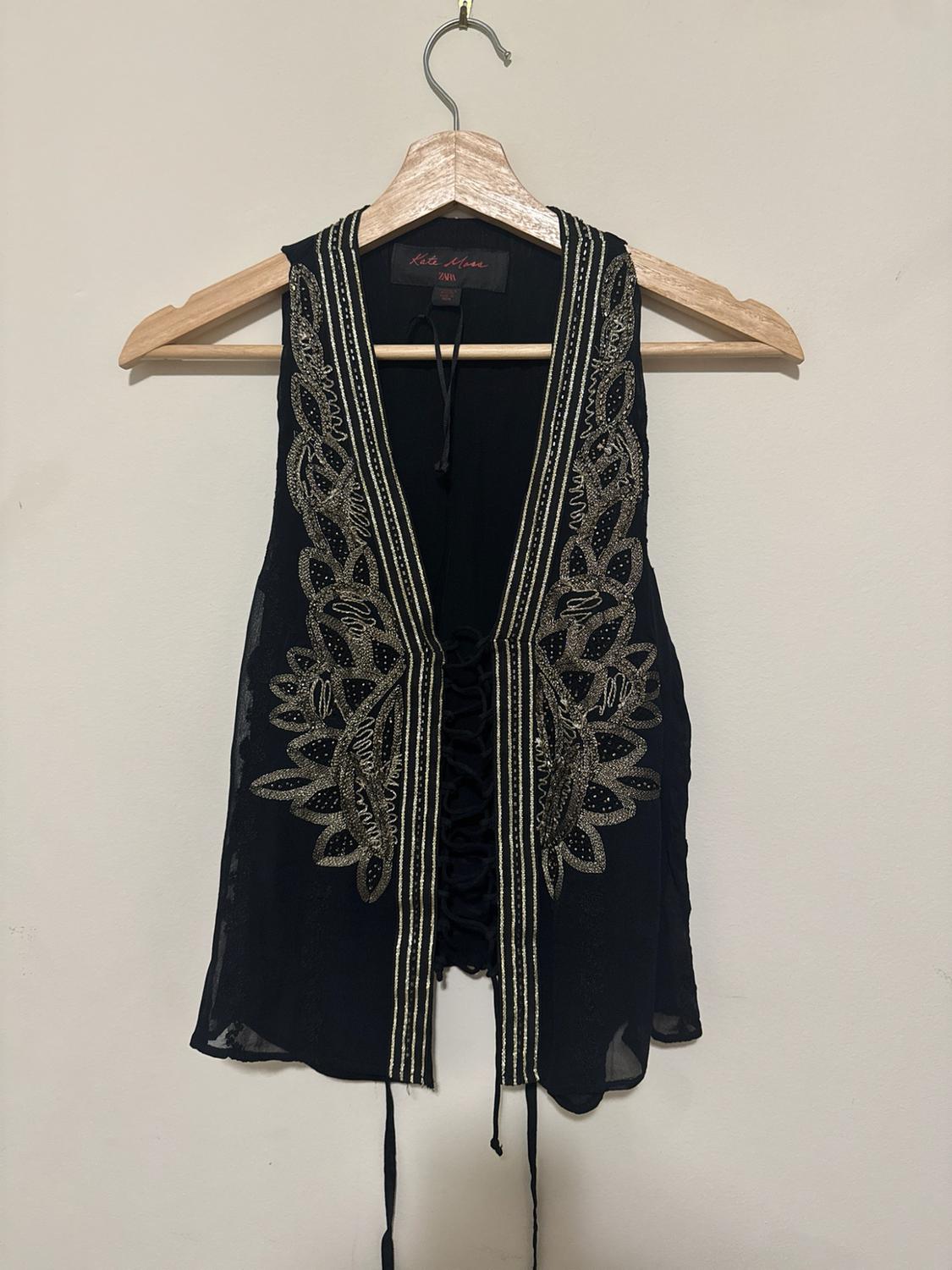 Black semi-sheer vest with gold embroidery and laces, on a wooden hanger against a white wall. Label: Kate Moss ZARA.