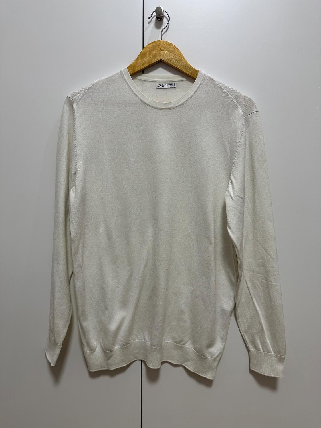 White, long-sleeved jumper hanging on a wooden hanger in front of a white wardrobe background. Visible label: "Zara".
