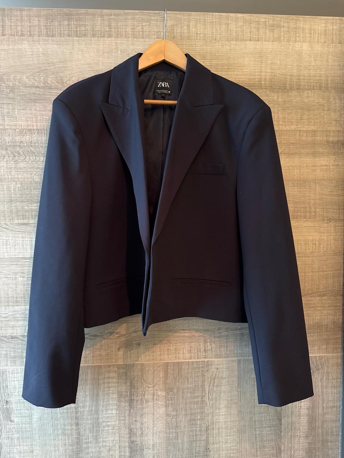 Black jacket hanging on a wooden hanger against a light wooden background. Visible label with text "ZARA".