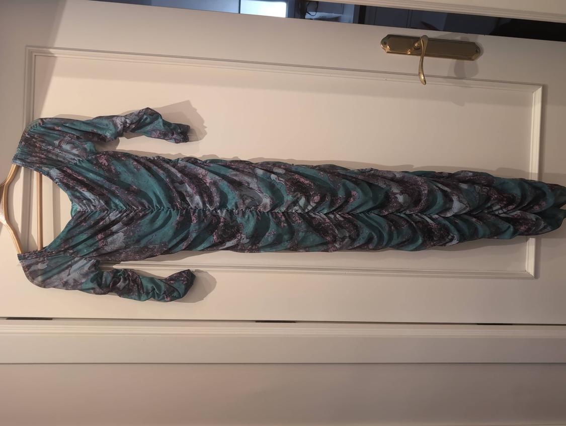 Long, ruched, long-sleeved dress with an abstract print in green and dark tones, hanging on a hanger over a white door.
