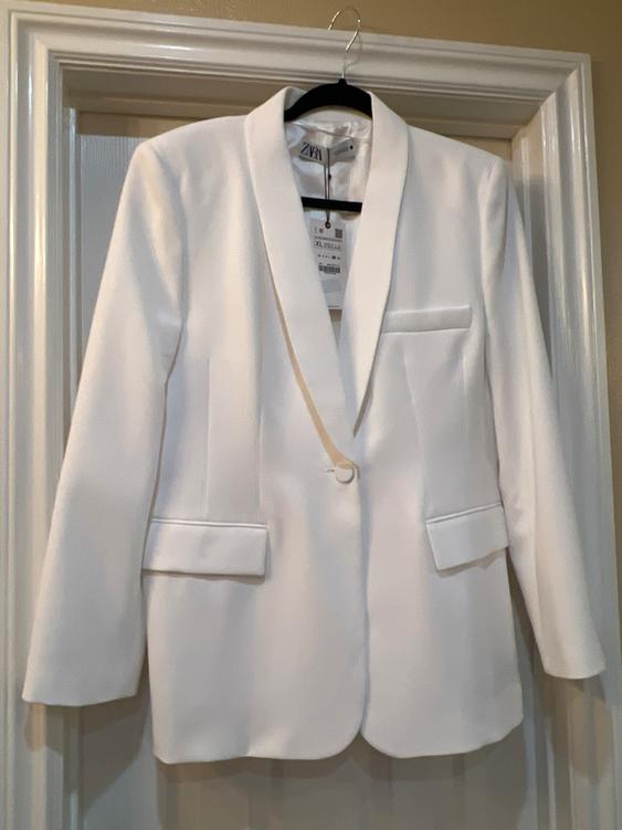 White blazer hanging on a black hanger over a door. Visible tag with text "ZARA" and size "XL".