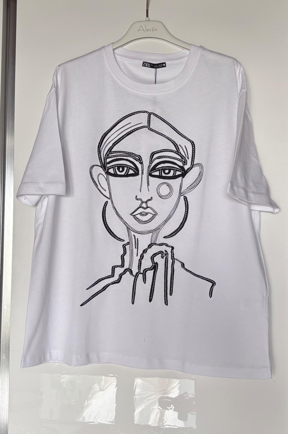 White T-shirt on a hanger with the text ‘Alesya’. Black line drawing of a woman’s face and a ‘ZARA’ label at the neck.