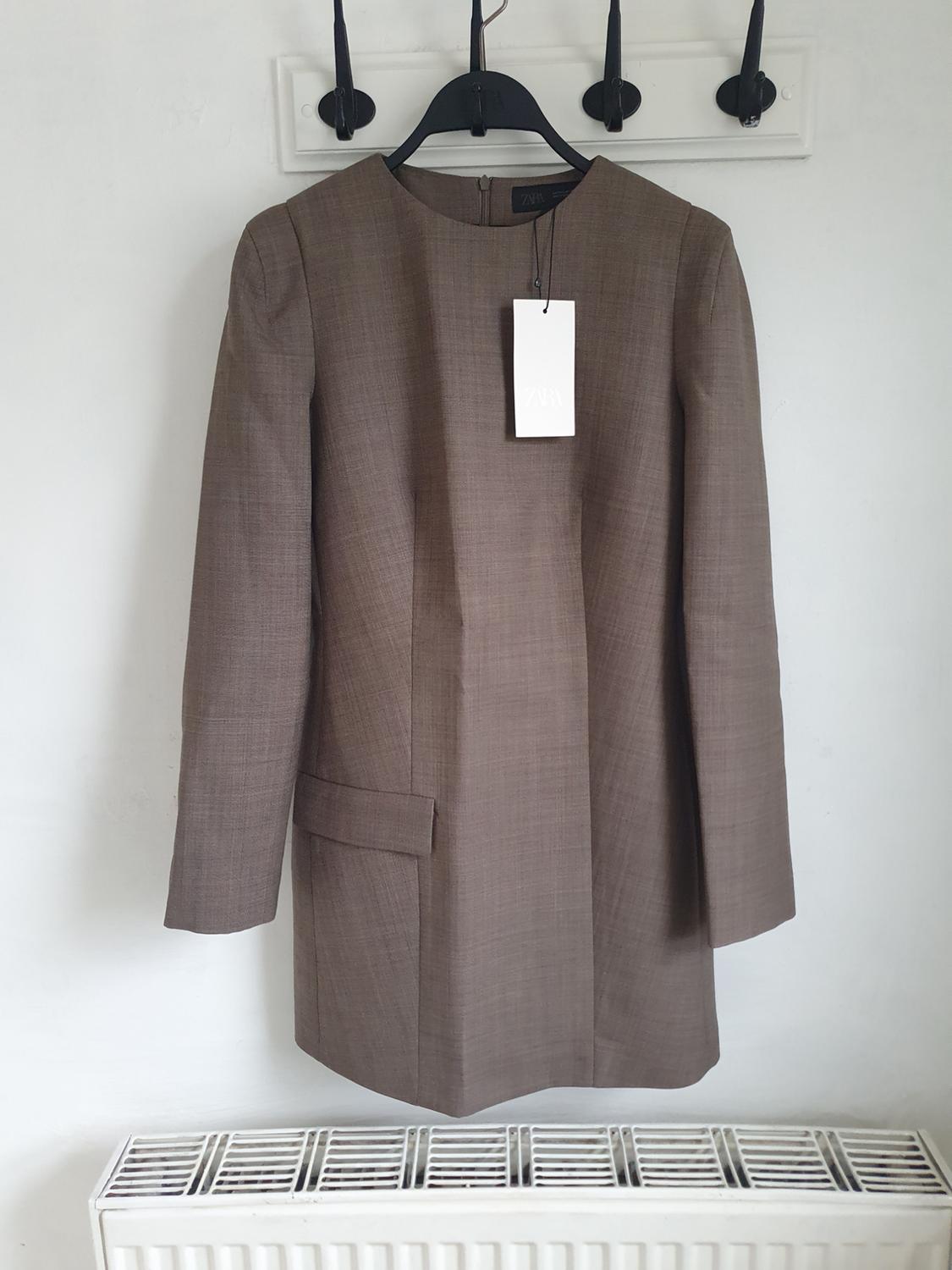 Brown coat hanging on a black hanger with a white Zara label, against a white background and a radiator underneath.