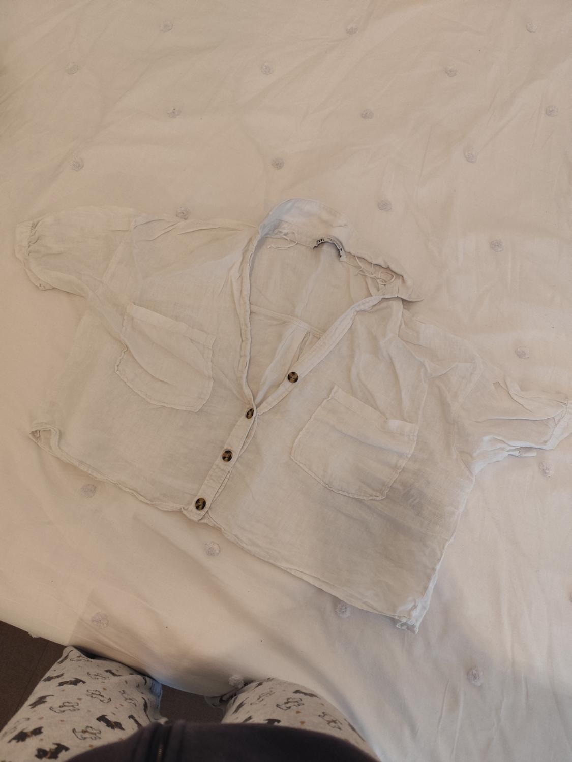 White short-sleeved shirt with brown buttons and two front pockets on a white quilt with embroidered details.