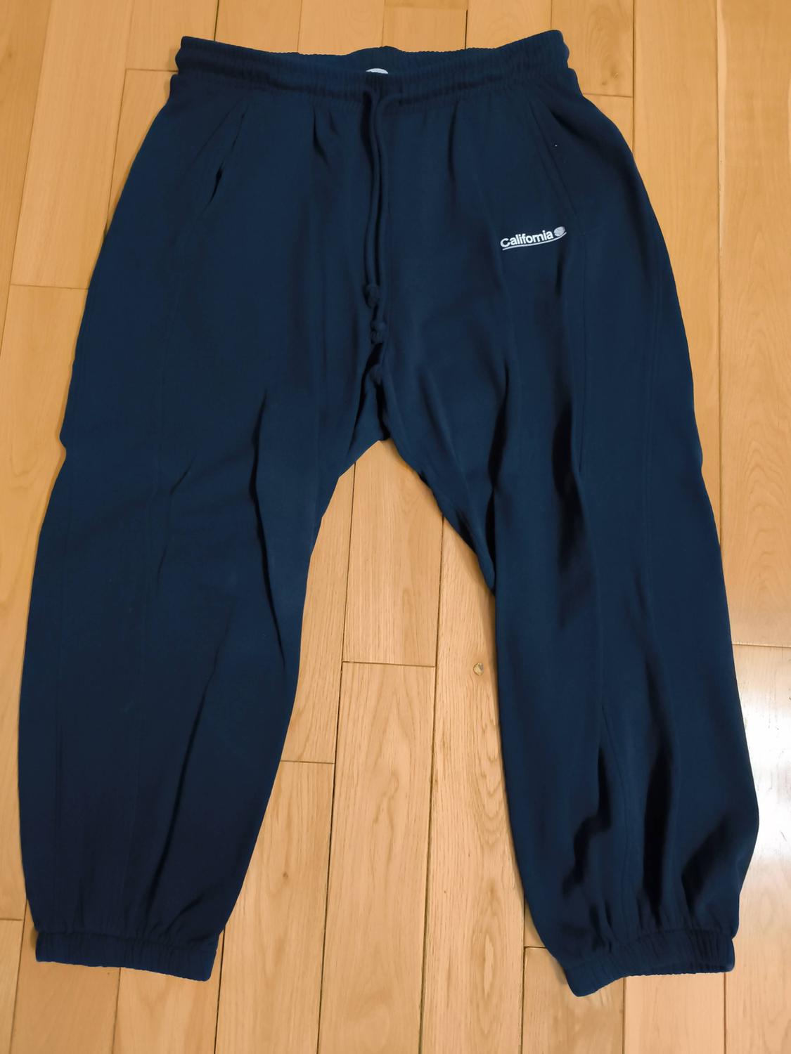 Dark blue sports trousers with an elasticated waist and 'California' logo on the left side, on a light wooden floor.