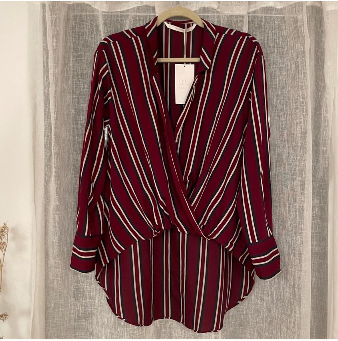 Vertical striped shirt in burgundy, white, and black tones, hanging on a hanger in front of a beige curtain.