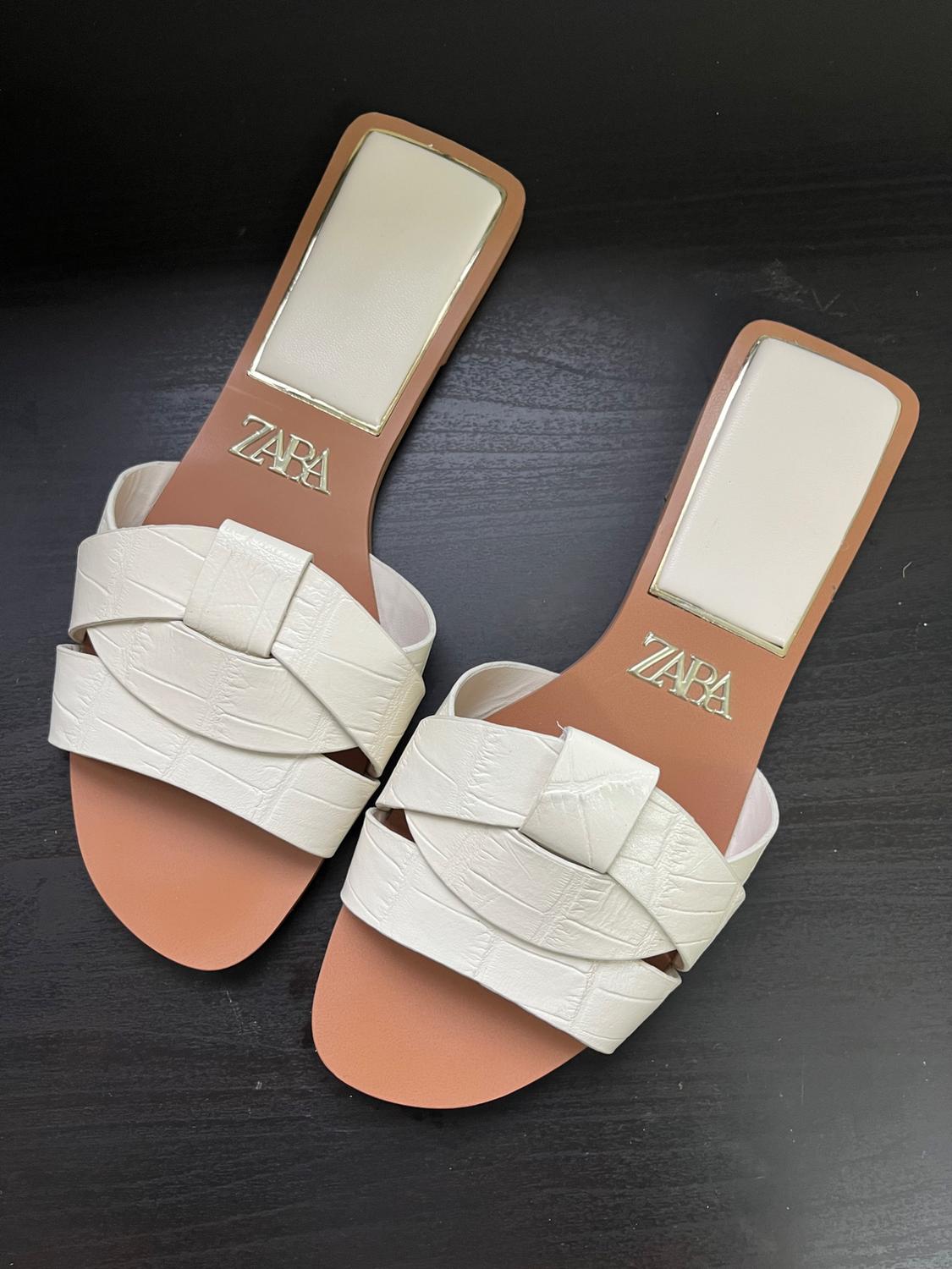 White flat sandals with interwoven straps on a black background. "ZARA" logo visible on the insole.